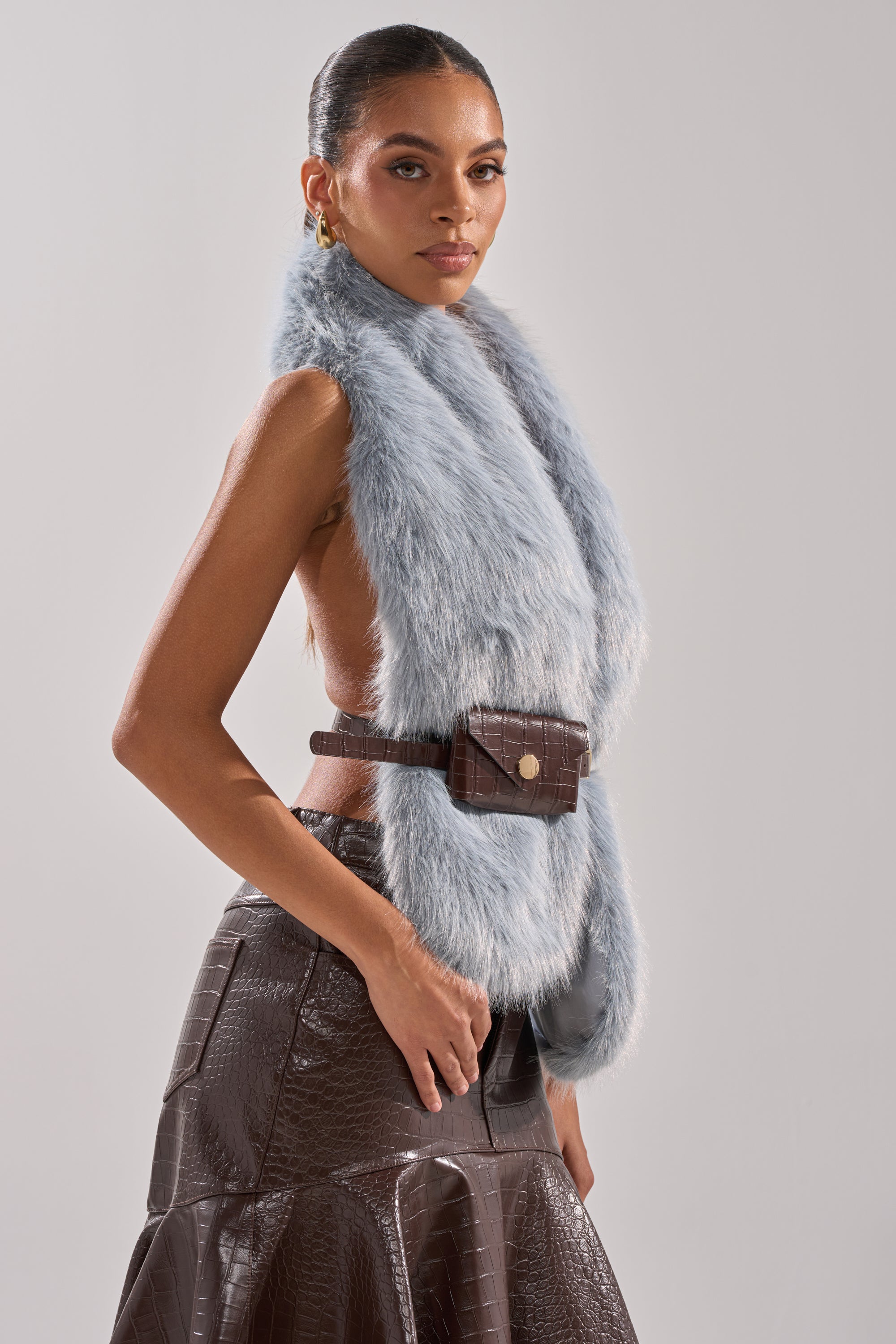 A woman poses in the ITS GIVING FUR BLOUSE, a sleeveless icy blue top, paired with a brown crocodile-textured leather skirt and matching belt bag. She accessorizes with gold earrings, her sleek hair pulled back, against a plain light background. 2