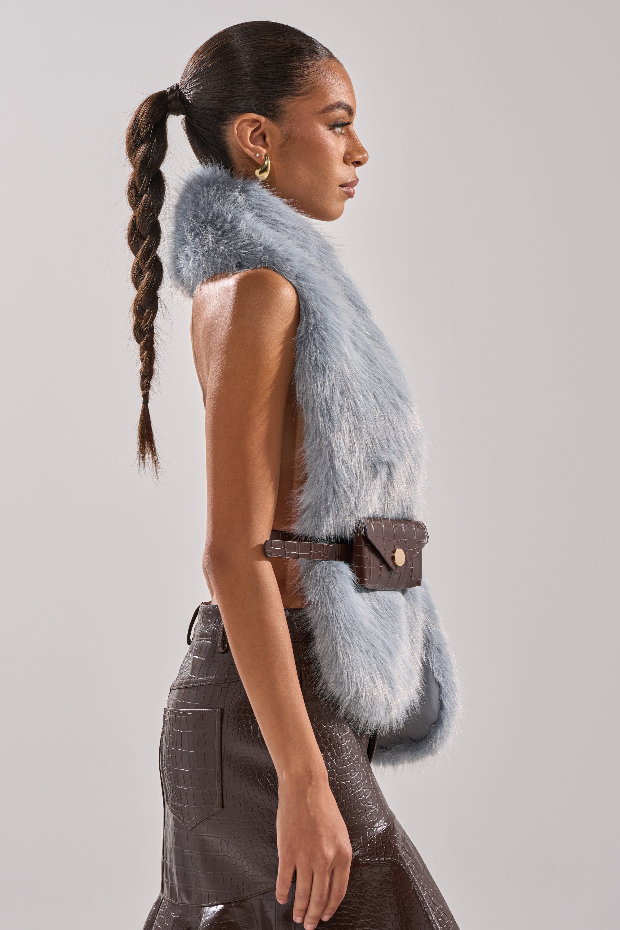 A woman with a long braided ponytail wears the sleeveless ITS GIVING FUR BLOUSE in light blue, styled with gold hoop earrings and high-waisted dark brown faux leather pants with an adjustable belt bag, standing in profile against a plain backdrop. 3