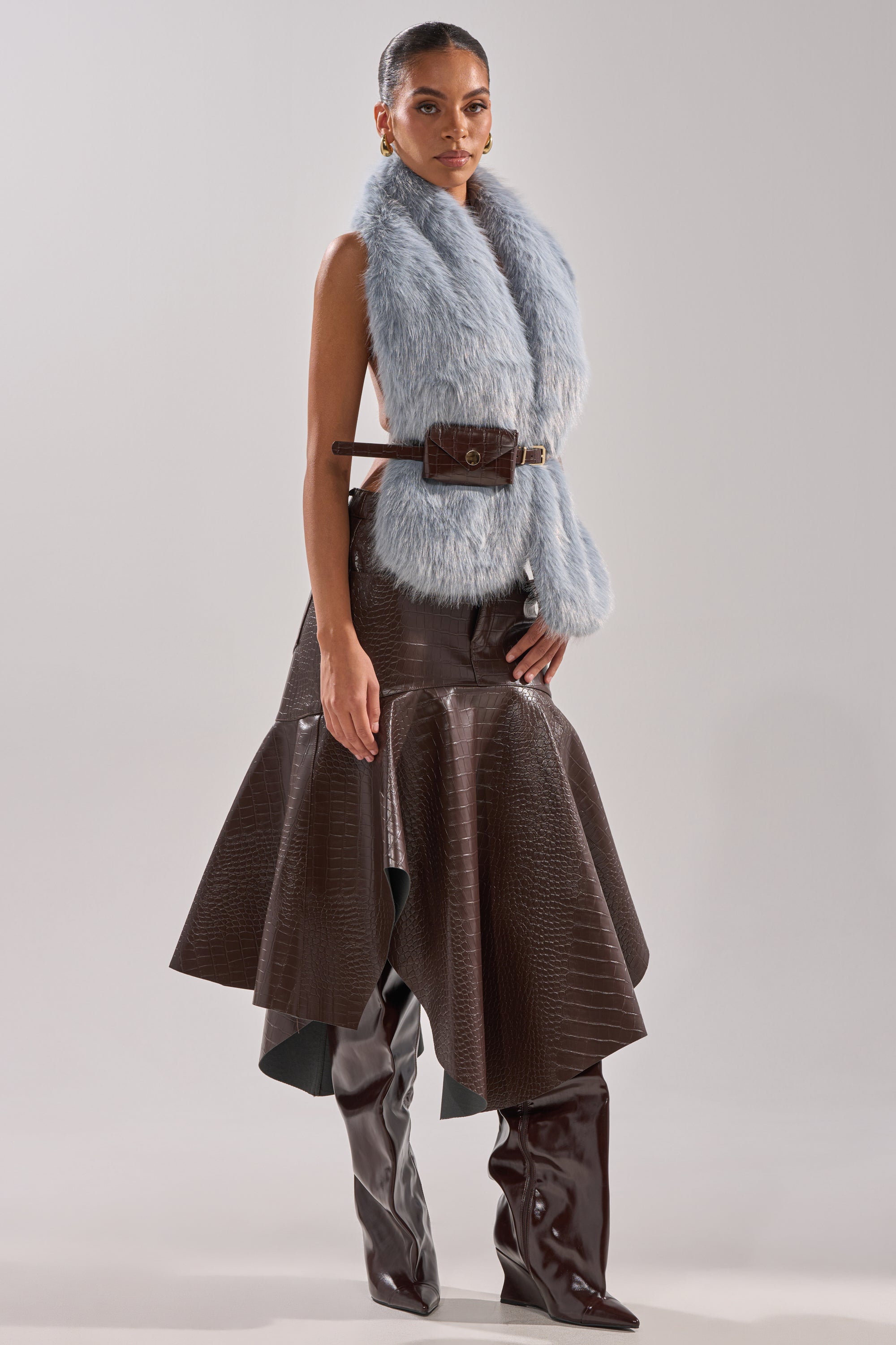 A woman poses in the ITS GIVING FUR BLOUSE styled with a brown textured asymmetrical wrap skirt, brown over-the-knee boots, and an adjustable belt bag against a plain light background. 5