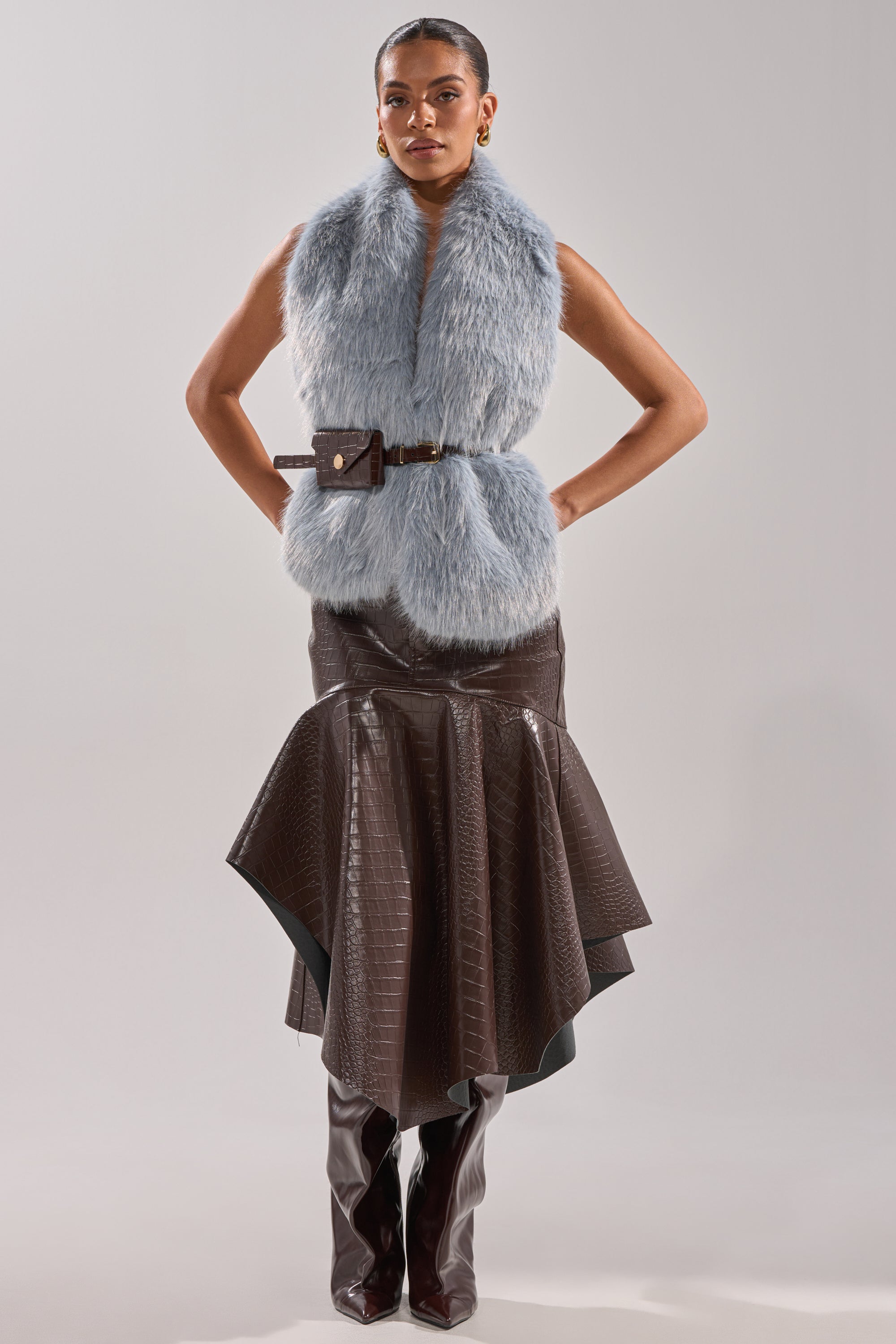 A woman poses against a plain background wearing the ITS GIVING FUR BLOUSE, a sleeveless light blue faux fur vest with an adjustable waist belt, paired with a dark brown textured asymmetrical midi skirt and brown knee-high boots. 6
