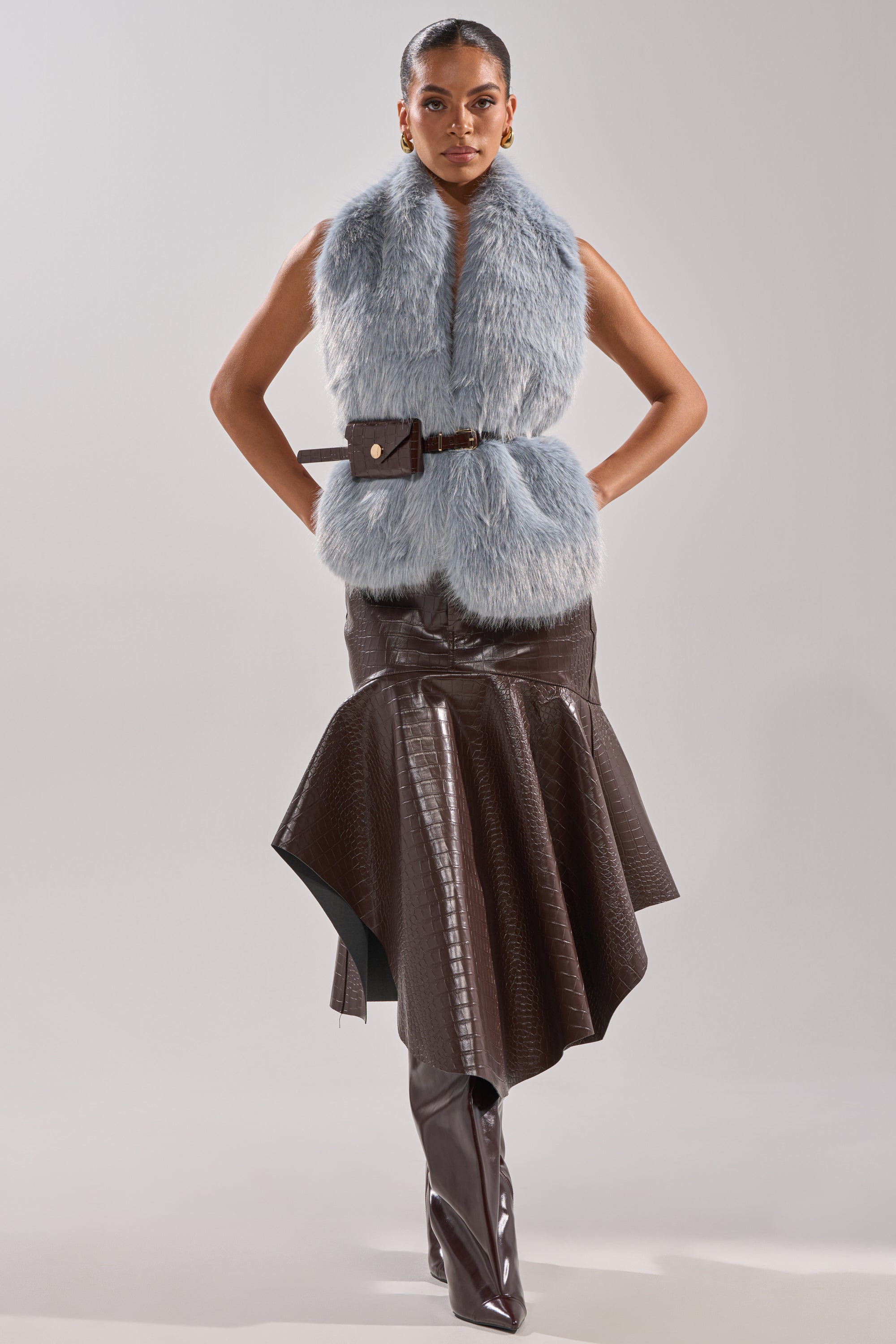 A woman stands confidently in the ITS GIVING FUR BLOUSE—light blue faux fur with an adjustable belt—paired with a textured dark brown asymmetrical skirt and tall matching brown boots against a plain background. 7