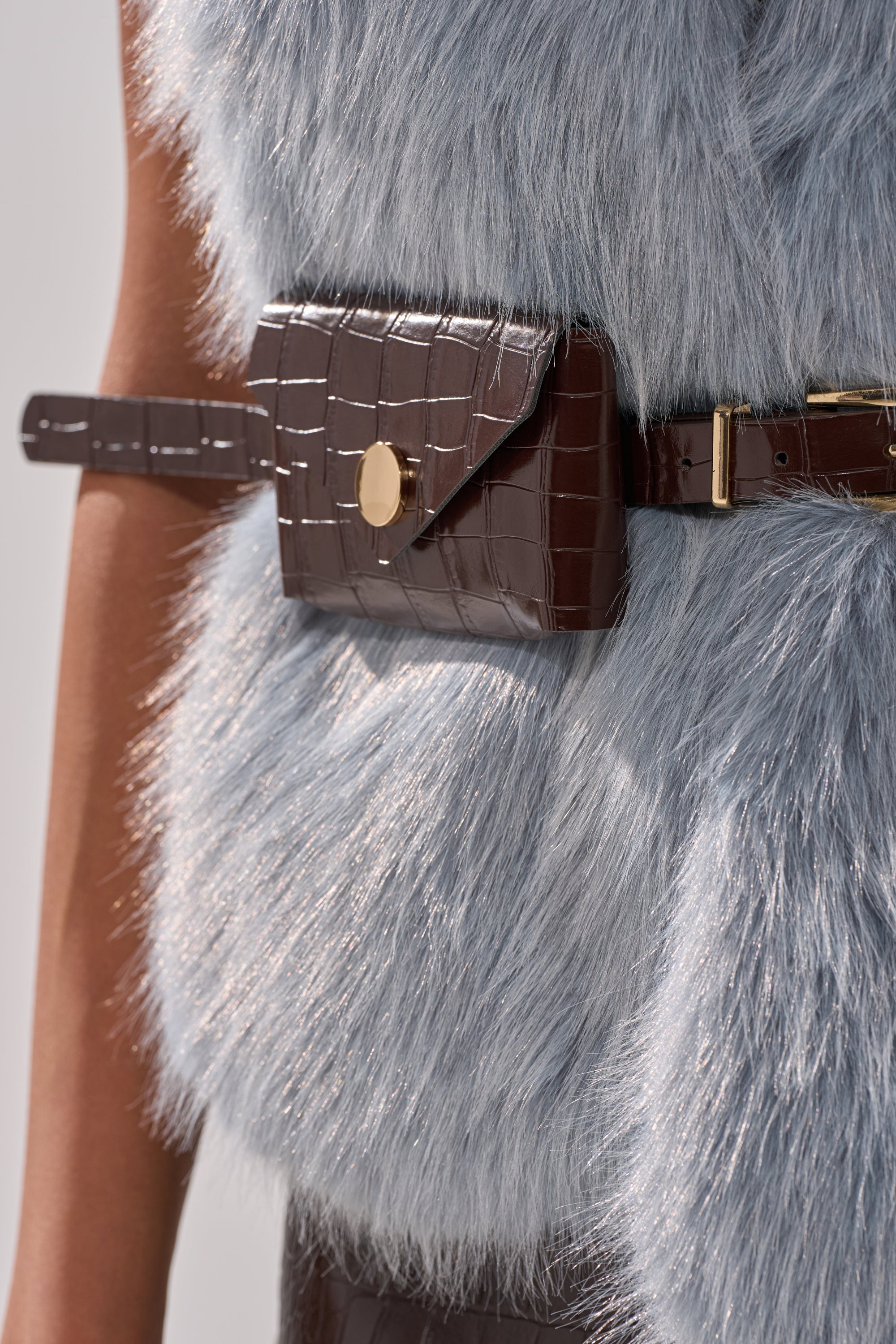 Close-up of a person in the ITS GIVING FUR BLOUSE, icy blue faux fur, styled with a dark brown crocodile-textured leather adjustable belt bag with a gold button closure. 8