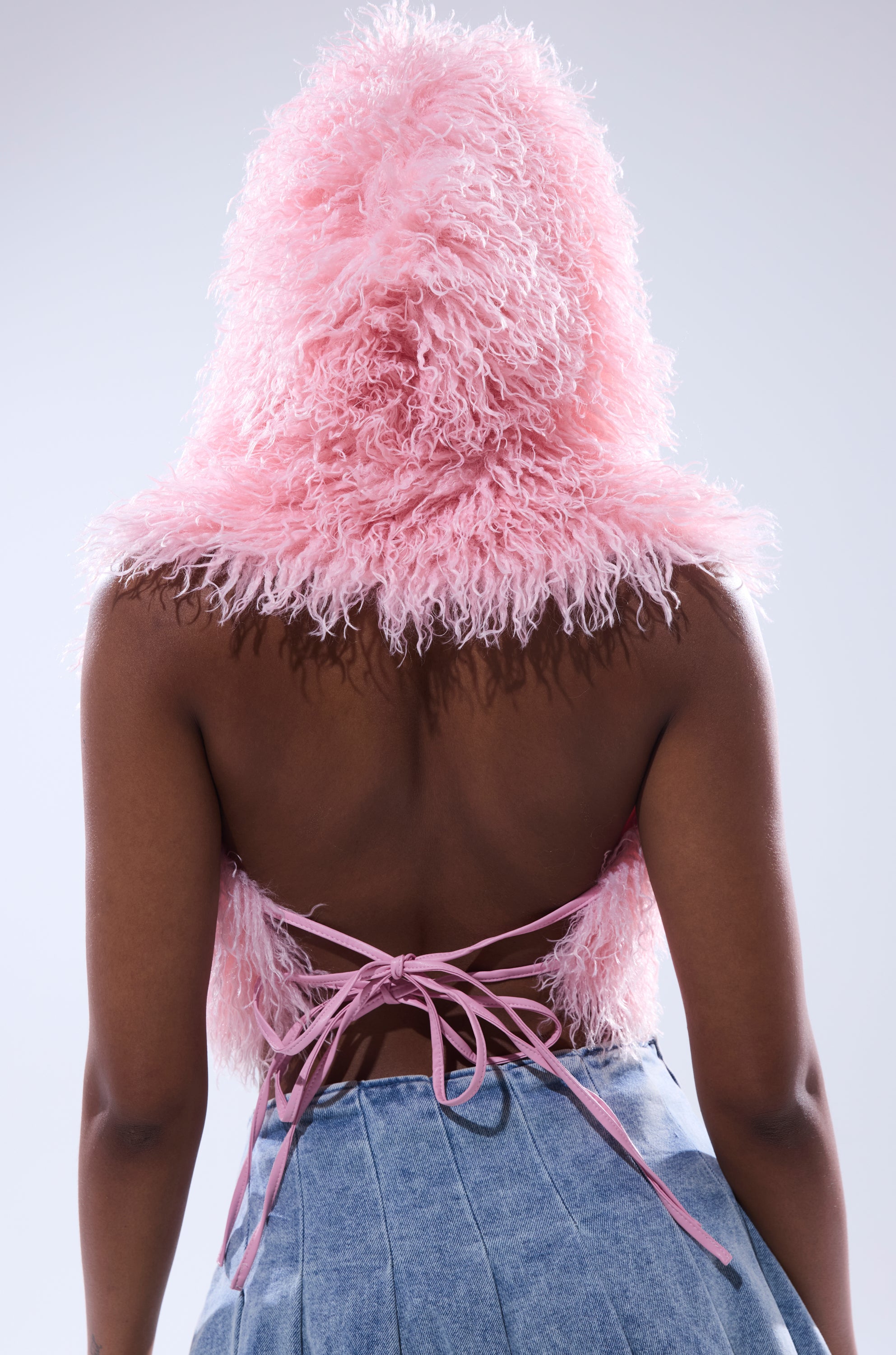 A person is shown from behind against a plain light background, wearing the THINK ABOUT YOU ALL THE TIME FUR BLOUSE IN PINK— a fuzzy, cropped top with an open back and lace-up ties—paired with high-waisted light blue denim pants. 5