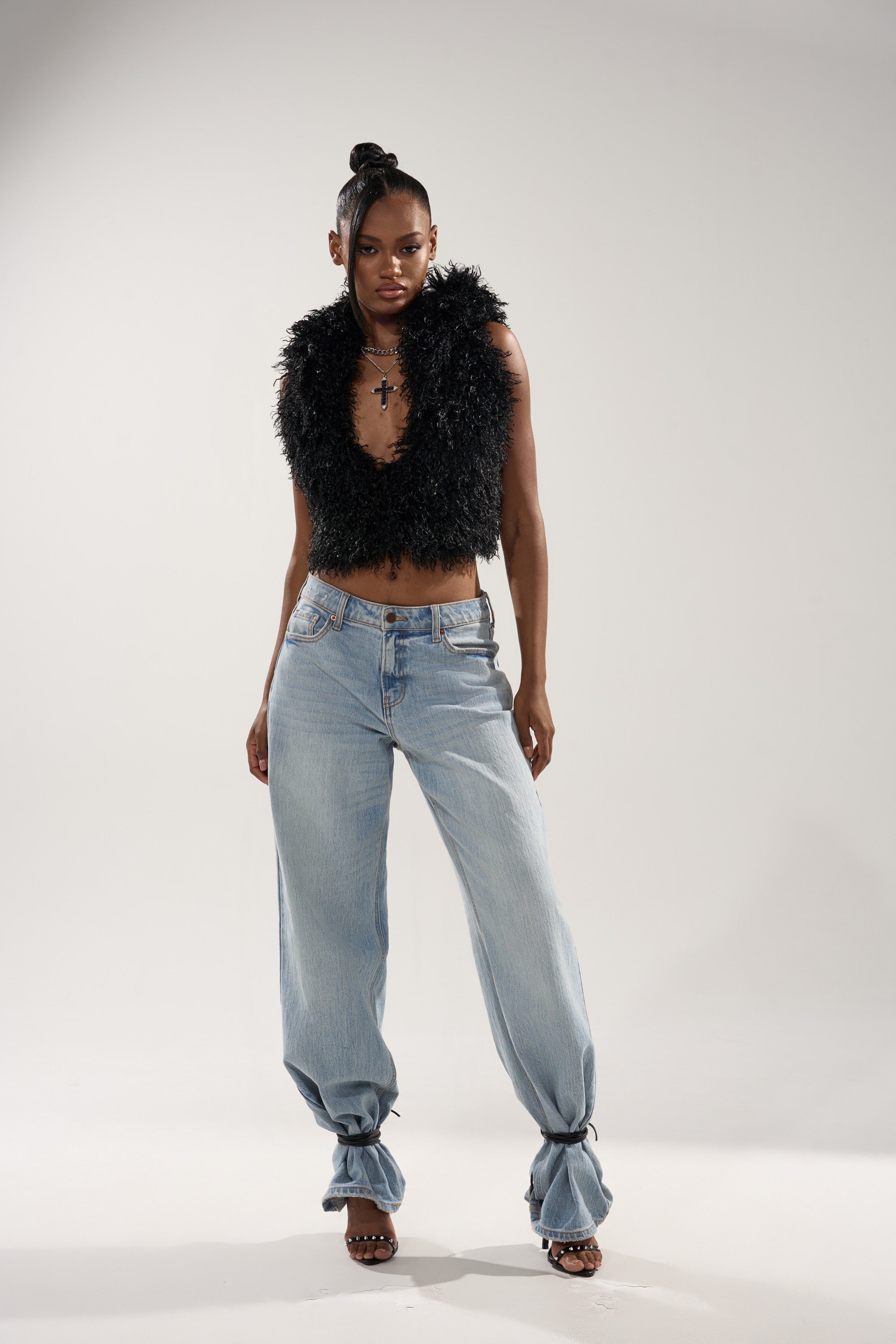 A woman stands confidently in the THINK ABOUT YOU ALL THE TIME FUR BLOUSE IN BLACK, paired with light blue baggy jeans, black heeled sandals, and layered necklaces against a plain light background.