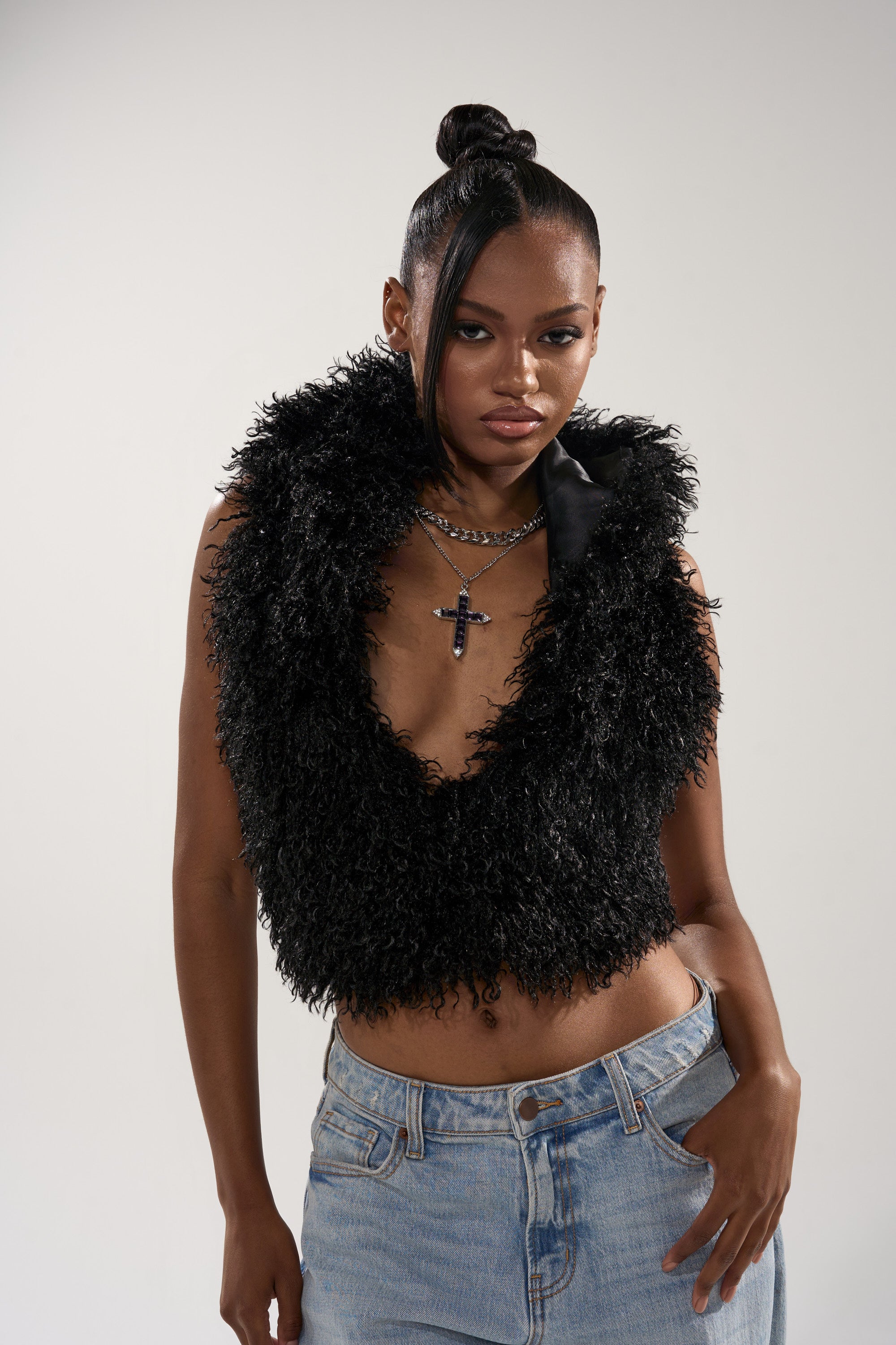 A woman confidently wears the THINK ABOUT YOU ALL THE TIME FUR BLOUSE IN BLACK, a sleeveless, hooded, deep V-neck top, paired with light blue jeans, layered necklaces, and a sleek updo with side-swept bangs against a plain background.