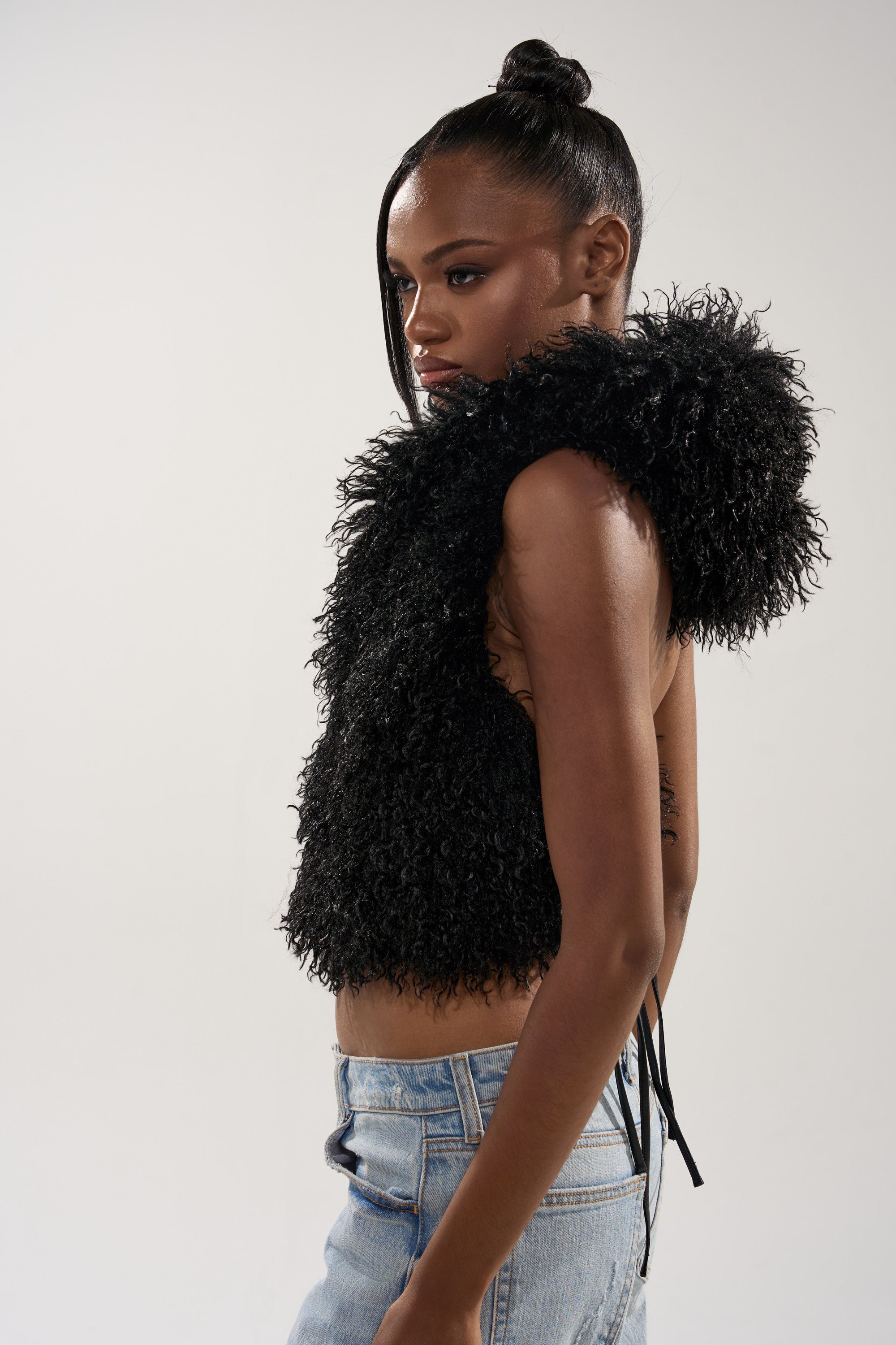 A woman with a high bun wears the THINK ABOUT YOU ALL THE TIME FUR BLOUSE IN BLACK, a sleeveless hooded blouse with large, fluffy shoulders, paired with light blue jeans while posing in profile against a plain white background.