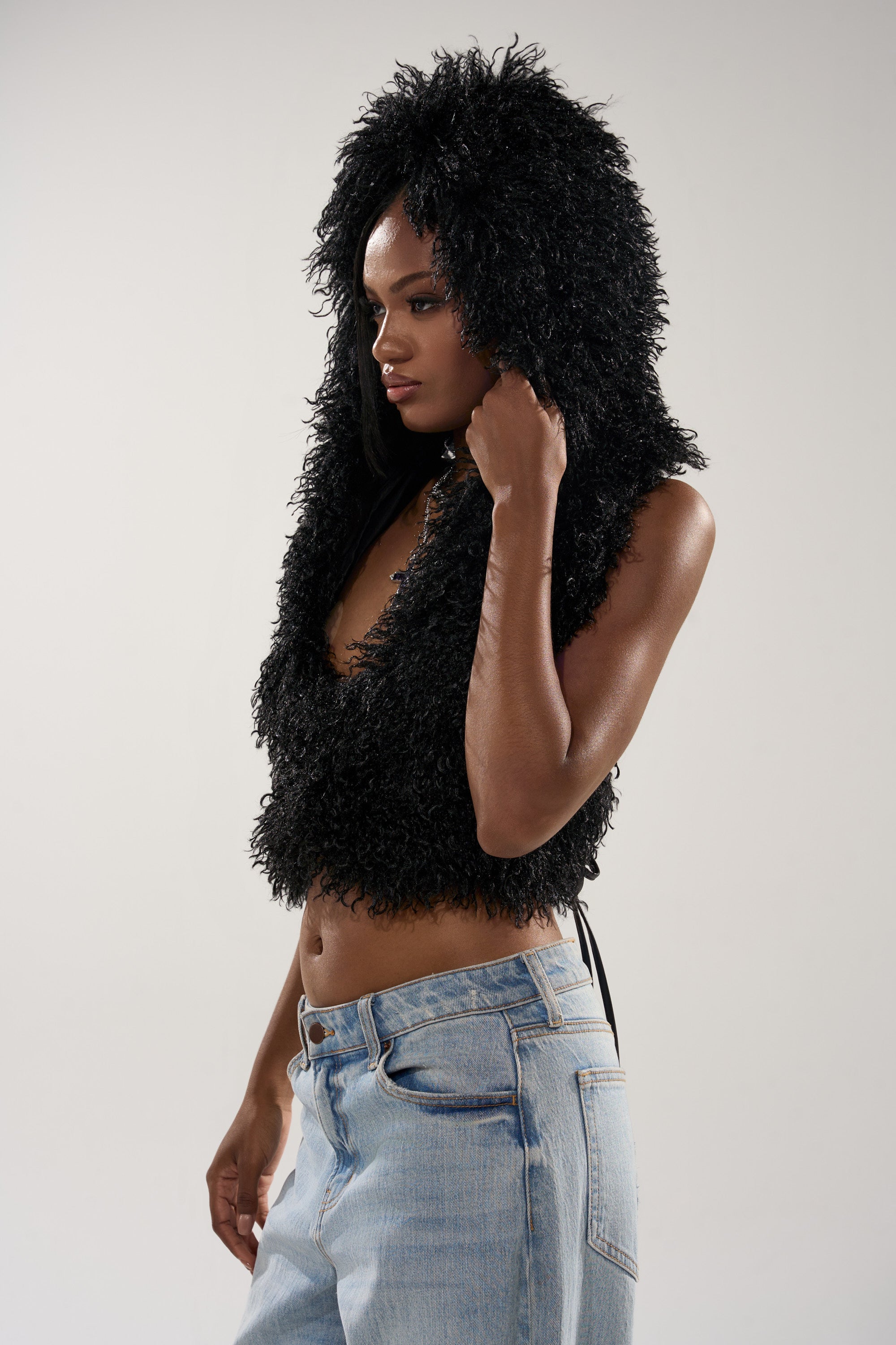 A woman in the THINK ABOUT YOU ALL THE TIME FUR BLOUSE IN BLACK, a cropped fur top with a hood, pairs it with light blue jeans and stands against a plain background, looking to the side and holding the edge of her hood.