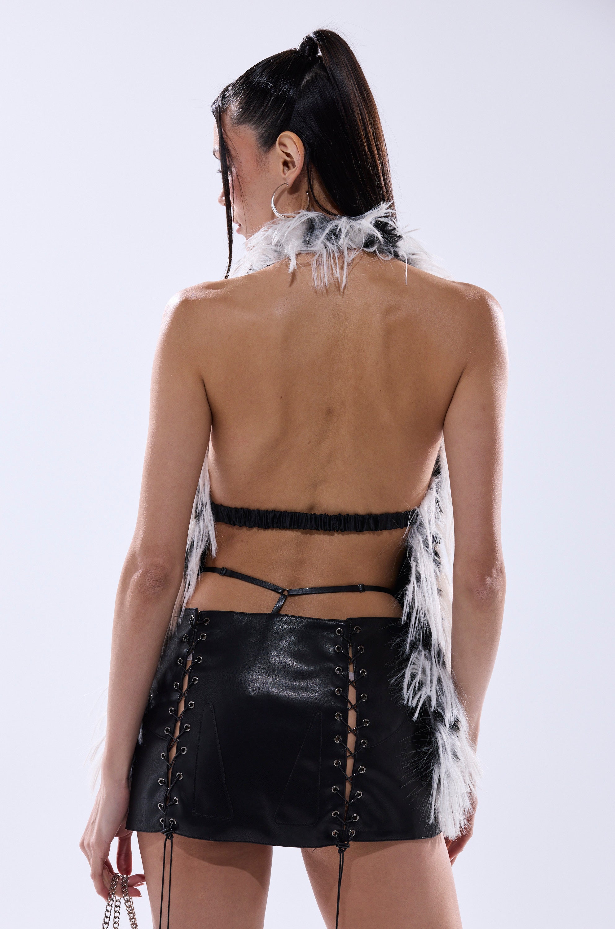 Wearing the ROUGHED UP FUR HALTER TOP, a woman with long dark hair in a high ponytail faces away, pairing the open-back faux fur halter with a black leather mini skirt and holding a chain strap accessory in her left hand. 4