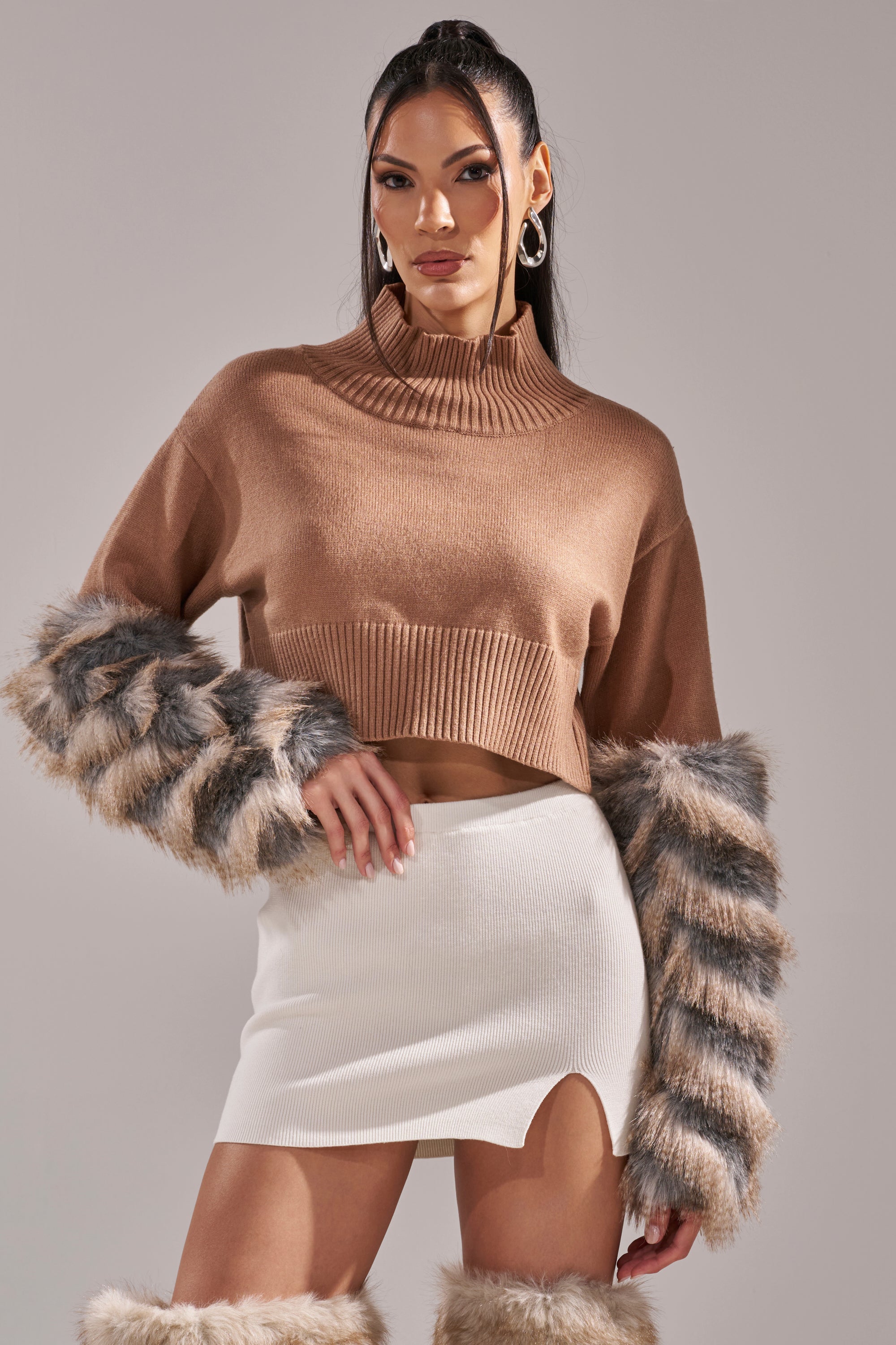 A woman models the PAWS OFF FUR CUFF SWEATER in tan with gray-striped faux fur accents, paired with a white front-slit mini skirt and matching furry boots. She wears her long dark hair in a high ponytail and large hoop earrings.