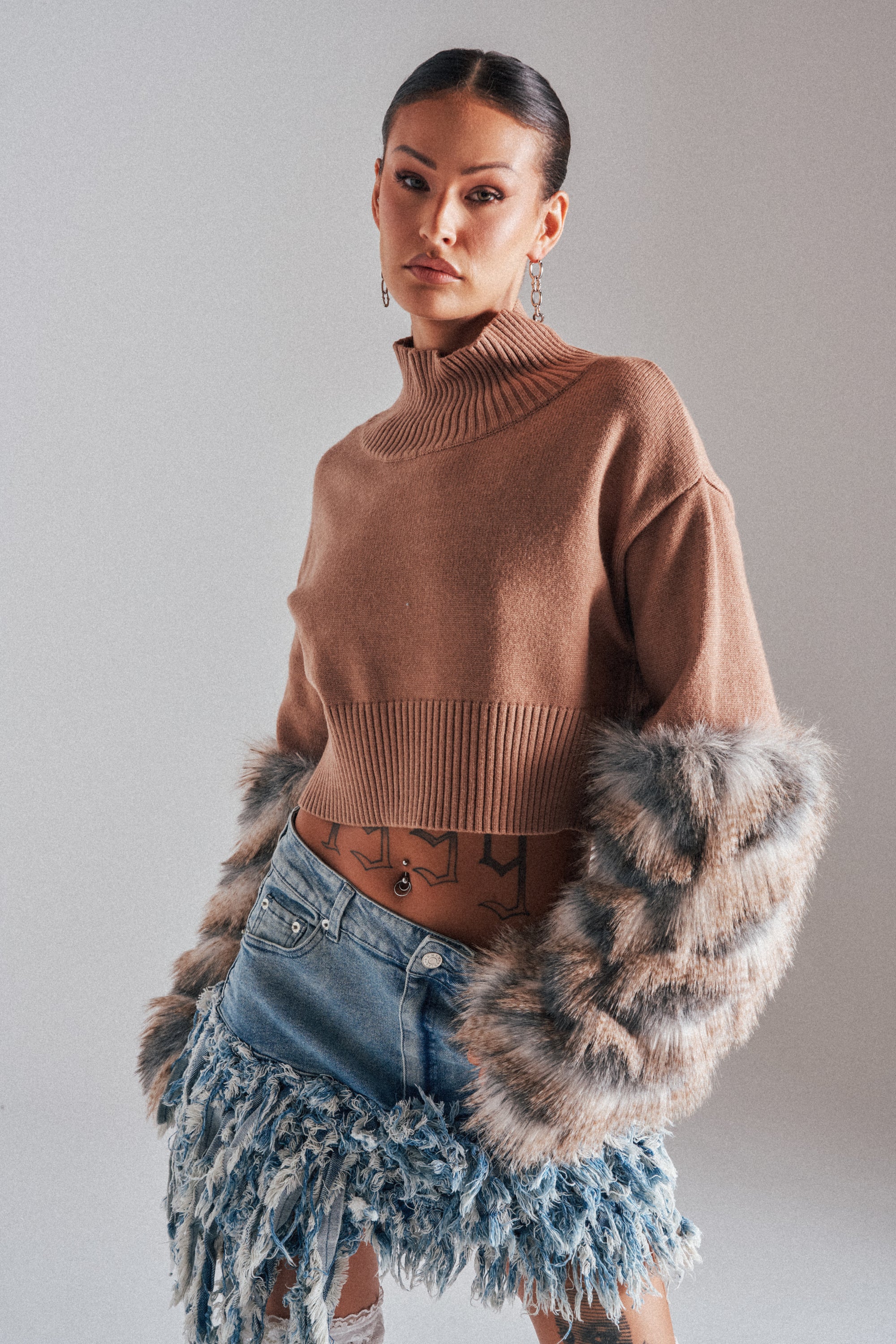 A woman models the PAWS OFF FUR CUFF SWEATER, featuring bold faux fur details, paired with a distressed fringed denim skirt. She accessorizes with long earrings, a belly button piercing, and showcases tattoos on her lower abdomen.