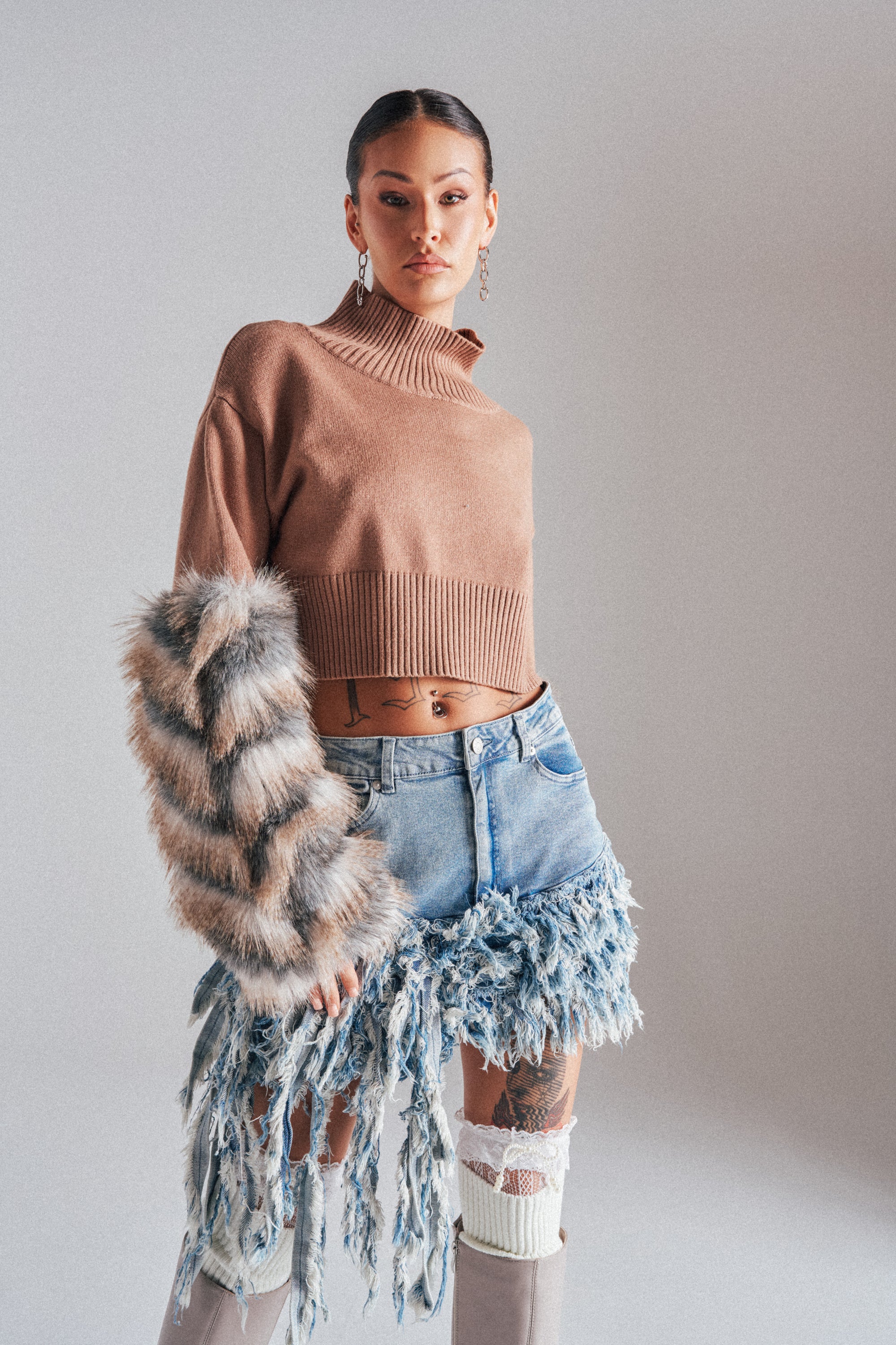 A woman models the PAWS OFF FUR CUFF SWEATER paired with a distressed denim skirt, knee-high beige boots, and hoop earrings, standing against a plain gray background.