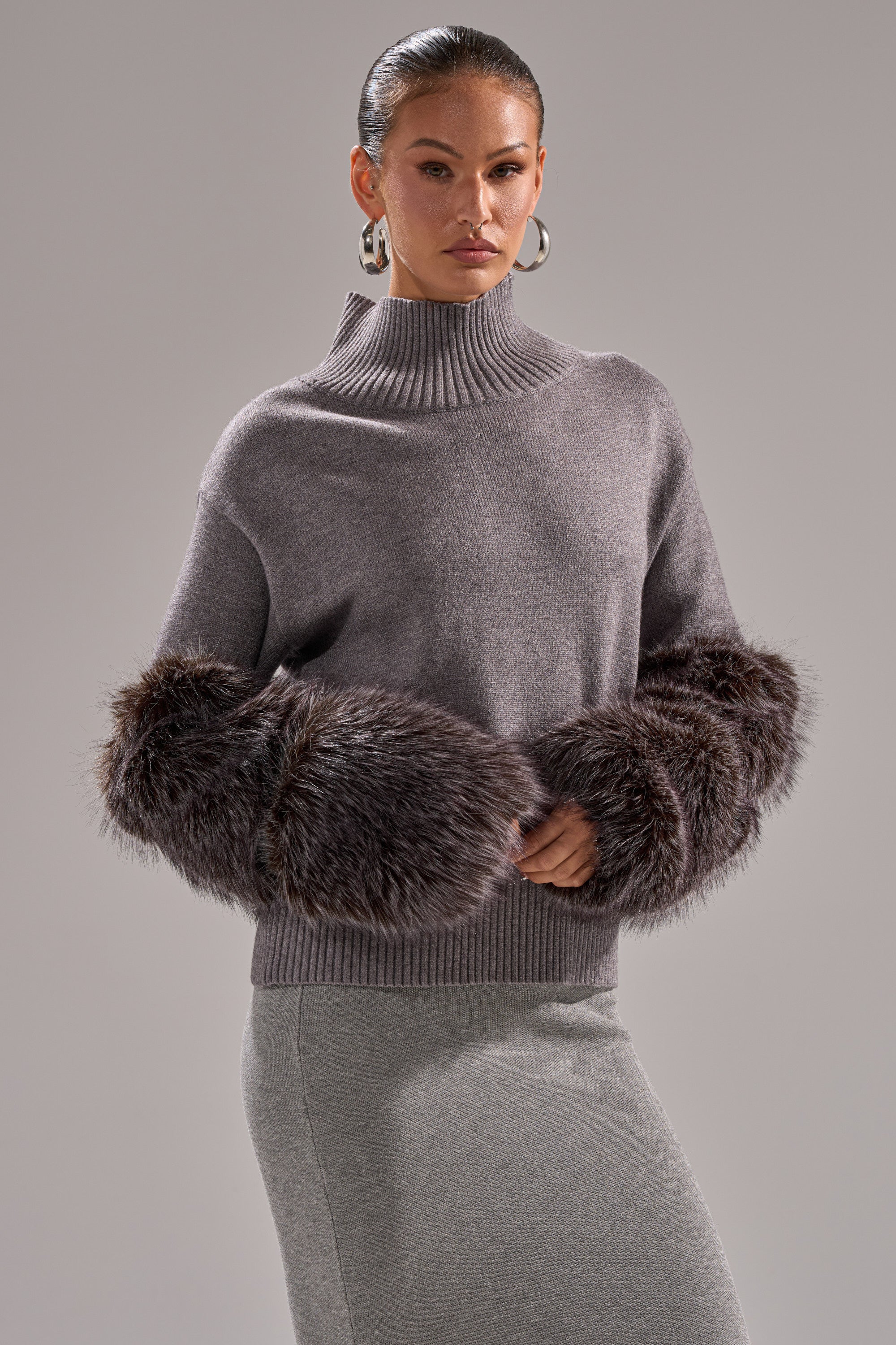 A woman in the TOUCH ME SWEATER WITH FUR CUFFS—a high-neck gray turtleneck with oversized dark faux fur cuffs—pairs it with a fitted gray skirt, large silver hoops, and slicked-back hair against a plain gray backdrop.