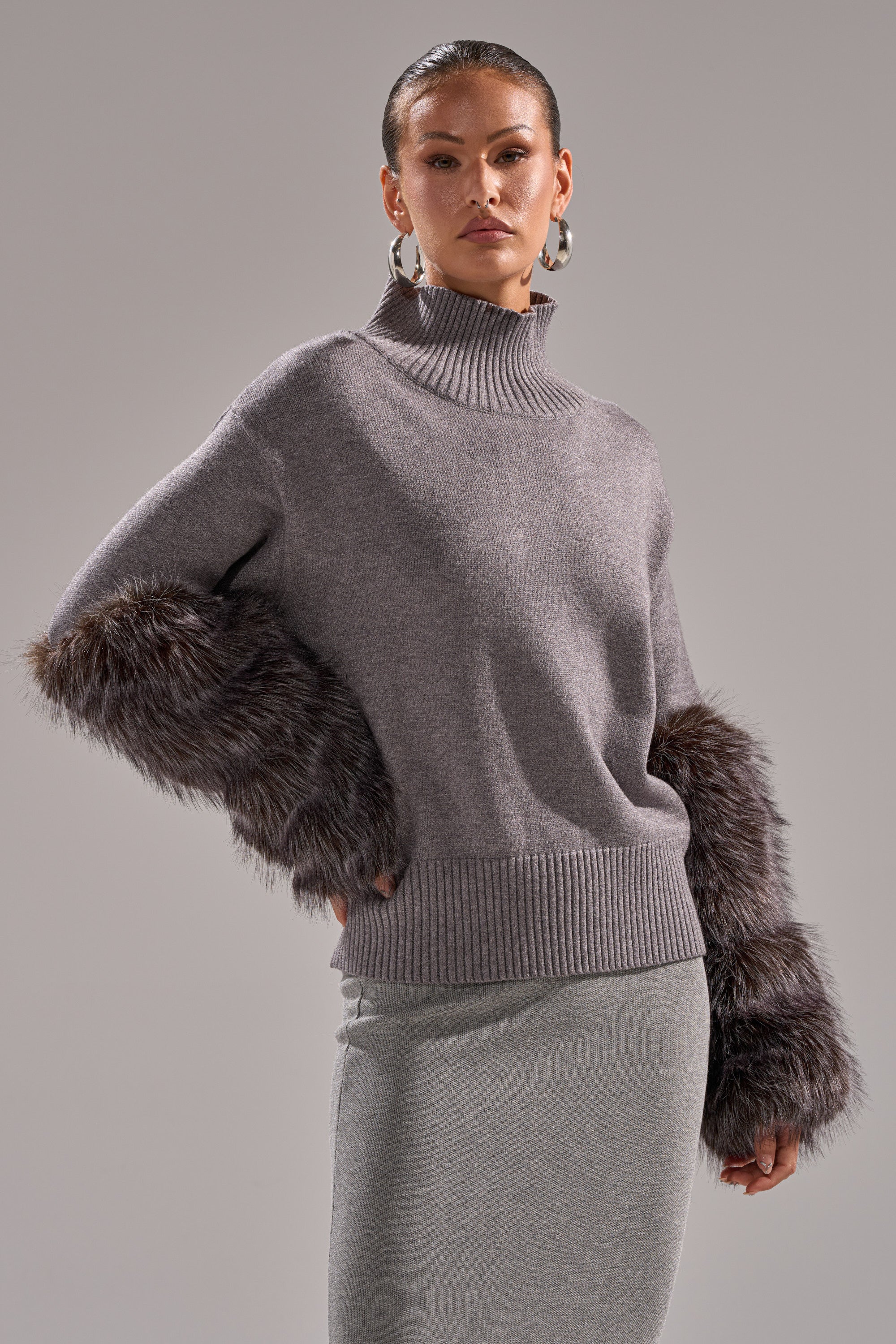 A woman wearing the TOUCH ME SWEATER WITH FUR CUFFS, a light gray turtleneck with chic fur cuffs, and a matching skirt poses against a neutral background with slicked-back hair, hoop earrings, and a confident expression.