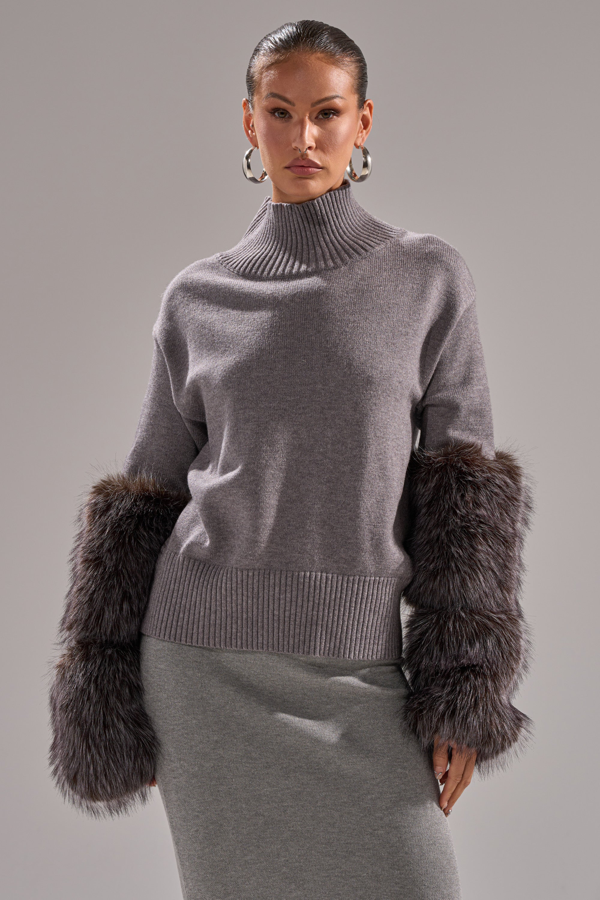 A woman models the TOUCH ME SWEATER WITH FUR CUFFS—a gray turtleneck featuring oversized dark fur cuffs—paired with a matching gray skirt, set against a plain gray background.