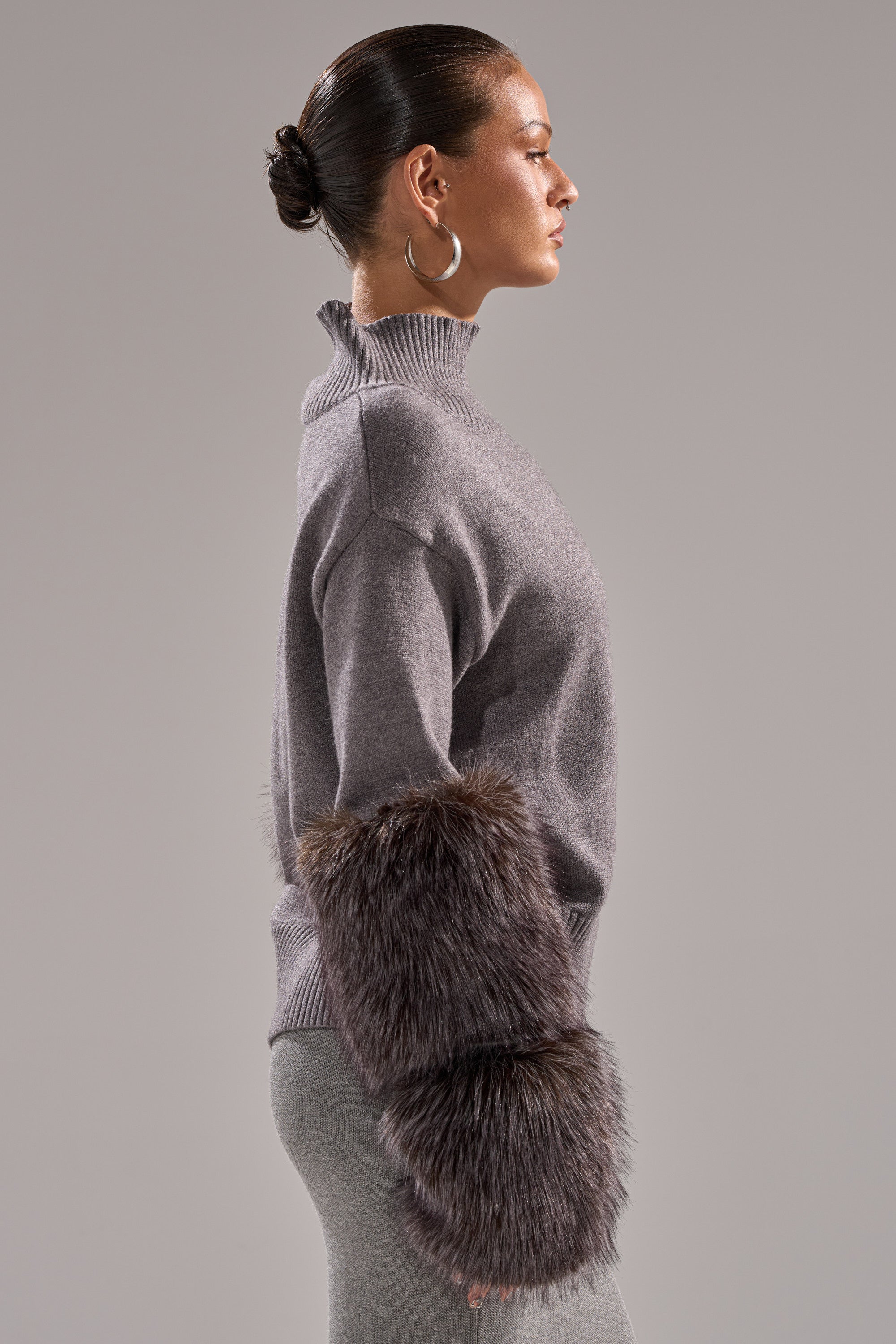 A woman with tied-back hair and silver hoop earrings models the TOUCH ME SWEATER WITH FUR CUFFS and matching gray pants against a plain gray background. The sweater features large, plush fur cuffs for a stylish accent.