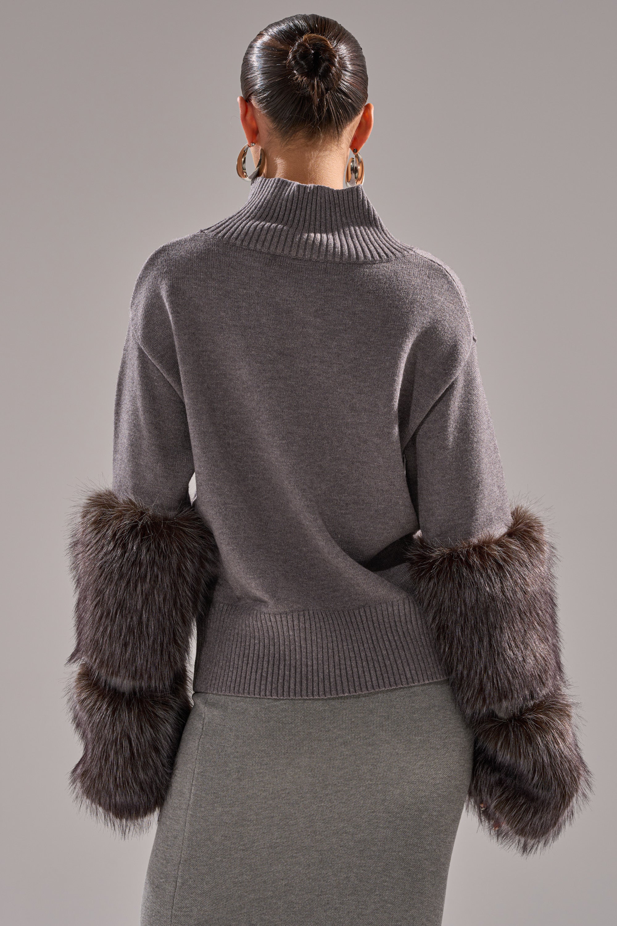 A woman faces away, wearing the TOUCH ME SWEATER WITH FUR CUFFS—gray with a wide ribbed collar, long sleeves, and large dark brown faux fur cuffs. Her hair is in a sleek bun and she accessorizes with large hoop earrings.