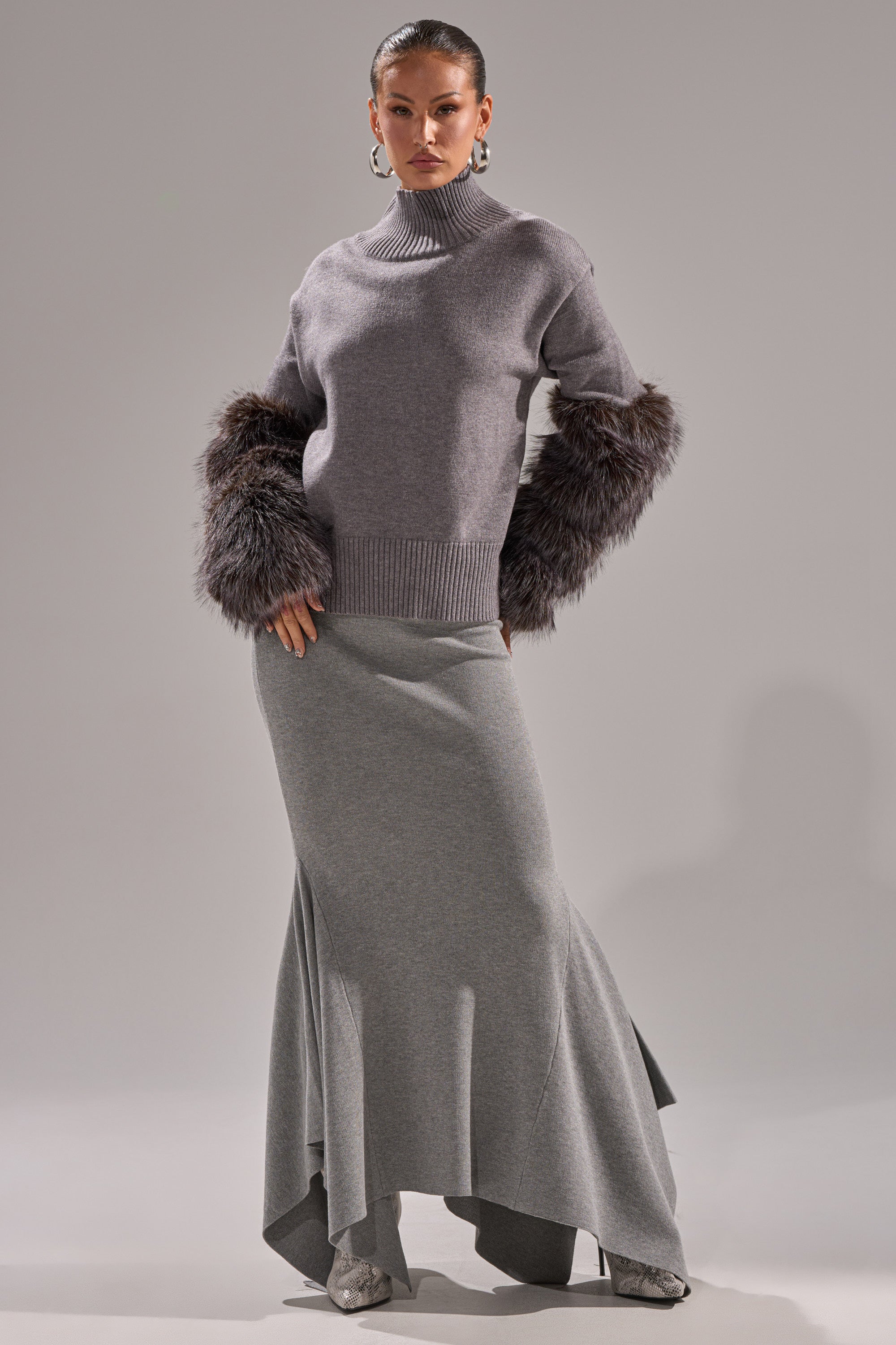 A woman stands confidently in a TOUCH ME SWEATER WITH FUR CUFFS, paired with a long grey asymmetric skirt, hoop earrings, and silver boots against a plain light grey background.