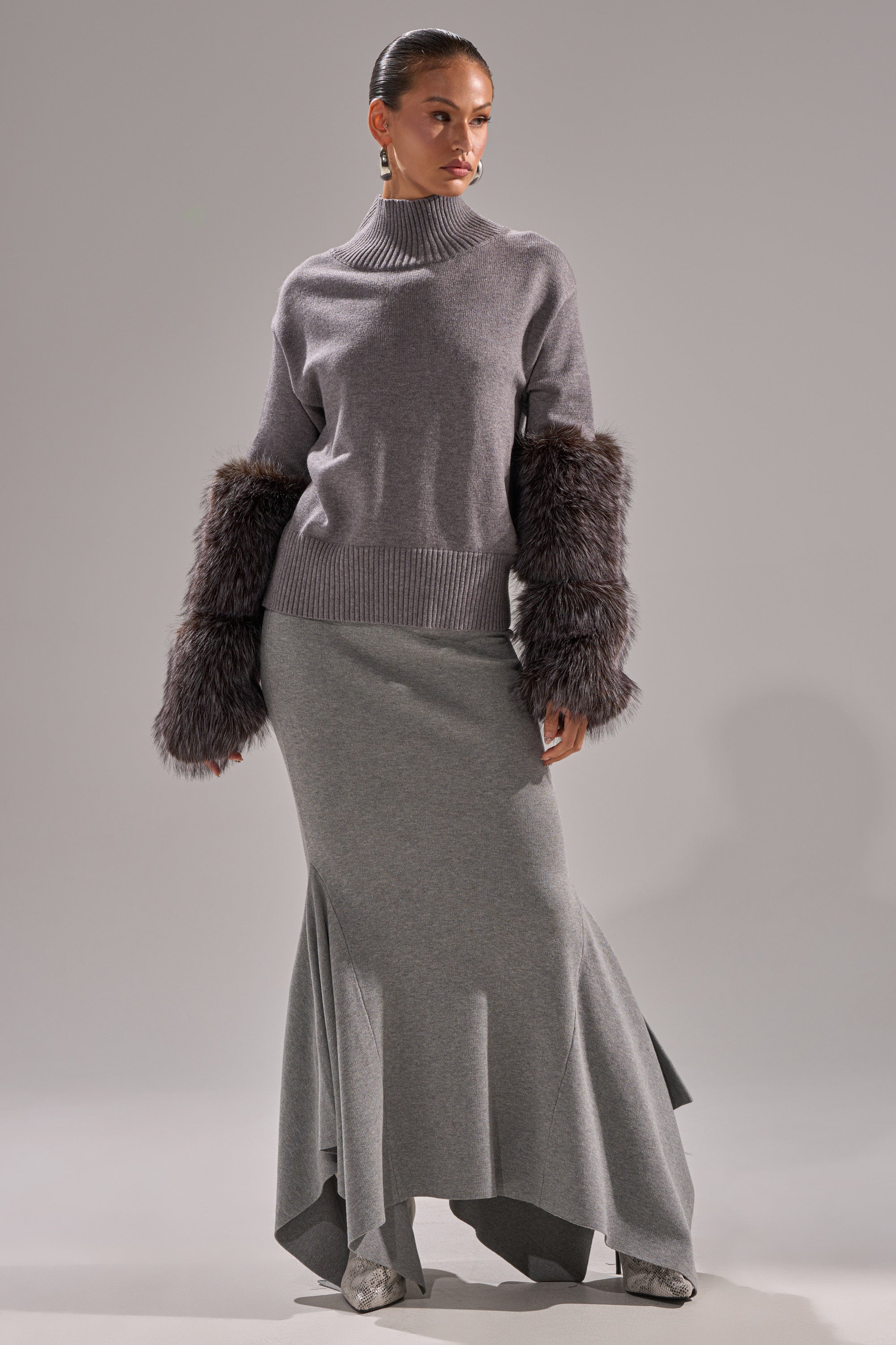 A woman poses in the TOUCH ME SWEATER WITH FUR CUFFS, a light gray turtleneck with dark gray fur cuffs, paired with a matching long skirt with a flared asymmetric hem, set against a plain light gray background.