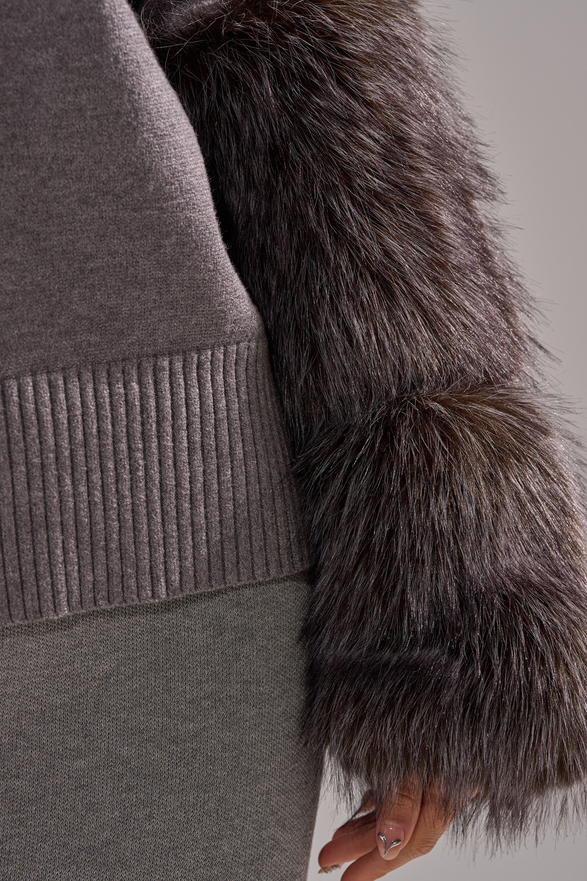 A close-up shows a person in the TOUCH ME SWEATER WITH FUR CUFFS, featuring a gray knit body, ribbed hem, and thick dark brown faux fur cuffs. Only part of the torso and one hand are visible.
