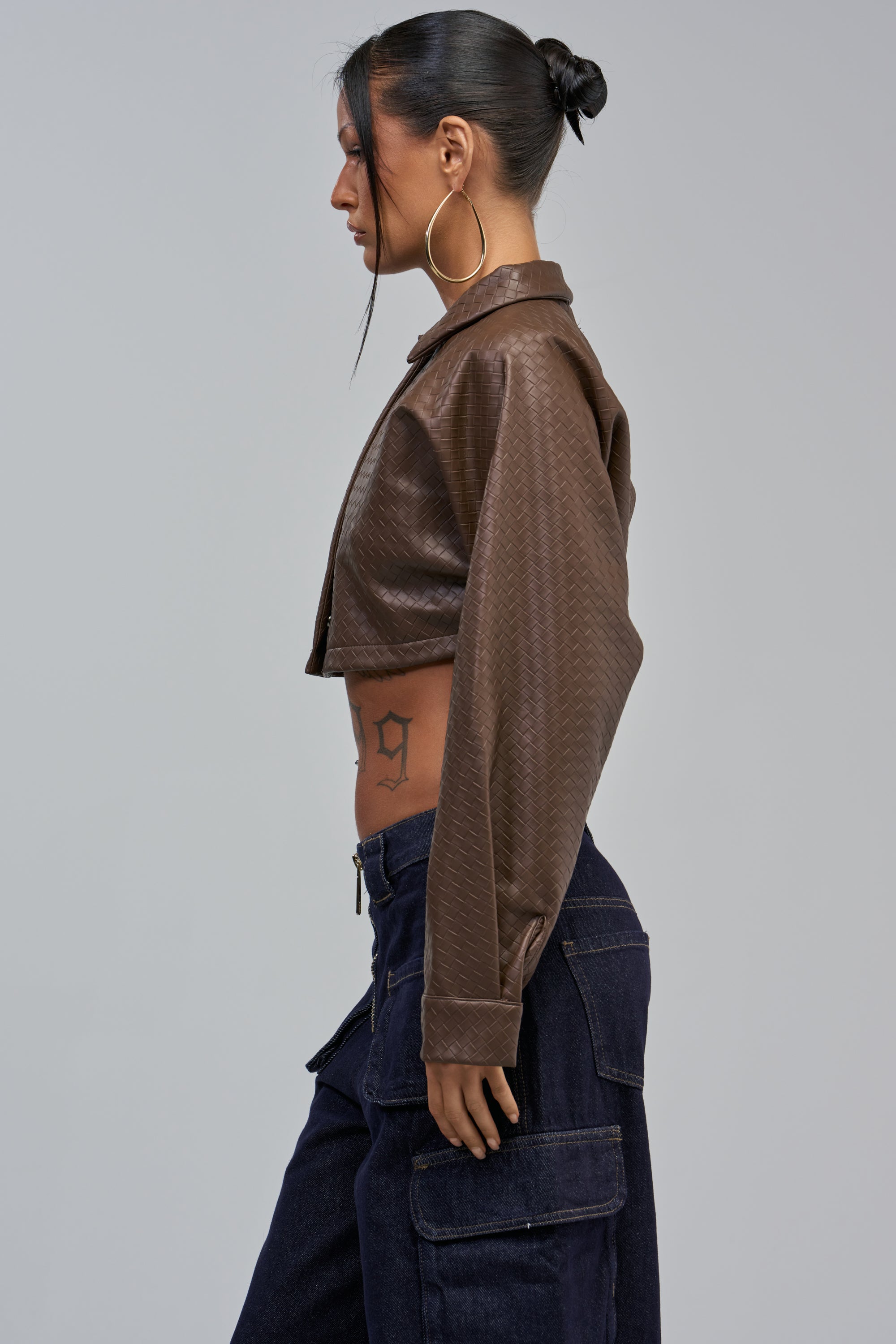 A person with a bun and large hoop earrings wears the LEILA WOVEN PU CROPPED BUTTON DOWN IN TAUPE with dark cargo pants, standing in profile against a plain gray background.