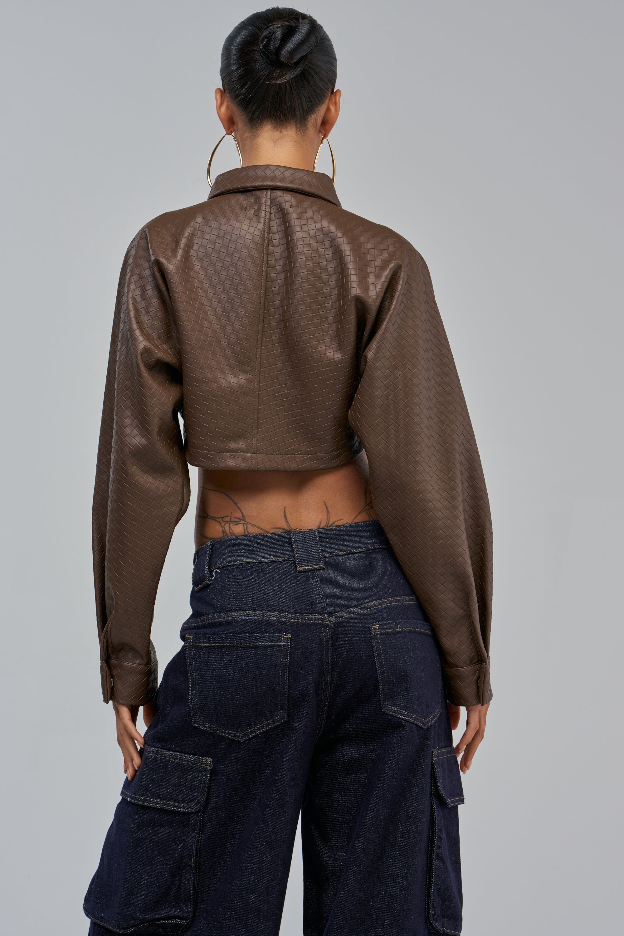 A person with a bun, hoop earrings, and lower back tattoos faces away, wearing the LEILA WOVEN PU CROPPED BUTTON DOWN IN TAUPE and dark blue cargo jeans.