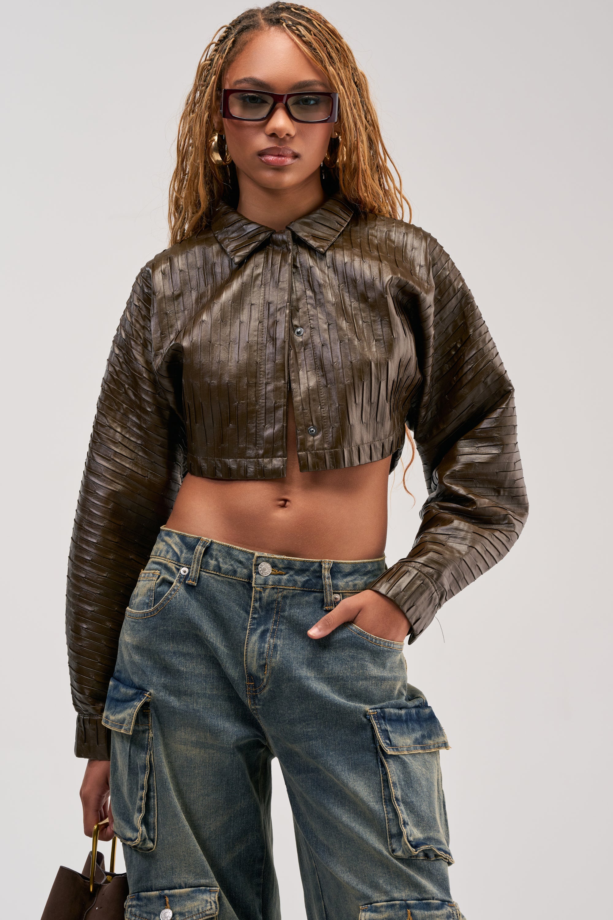 Wearing the JACQUELINE TEXTURED PU CROPPED BUTTON DOWN, paired with blue cargo jeans and sunglasses, a person poses confidently with one hand in their pocket against a plain background, capturing an effortless utility-chic style.