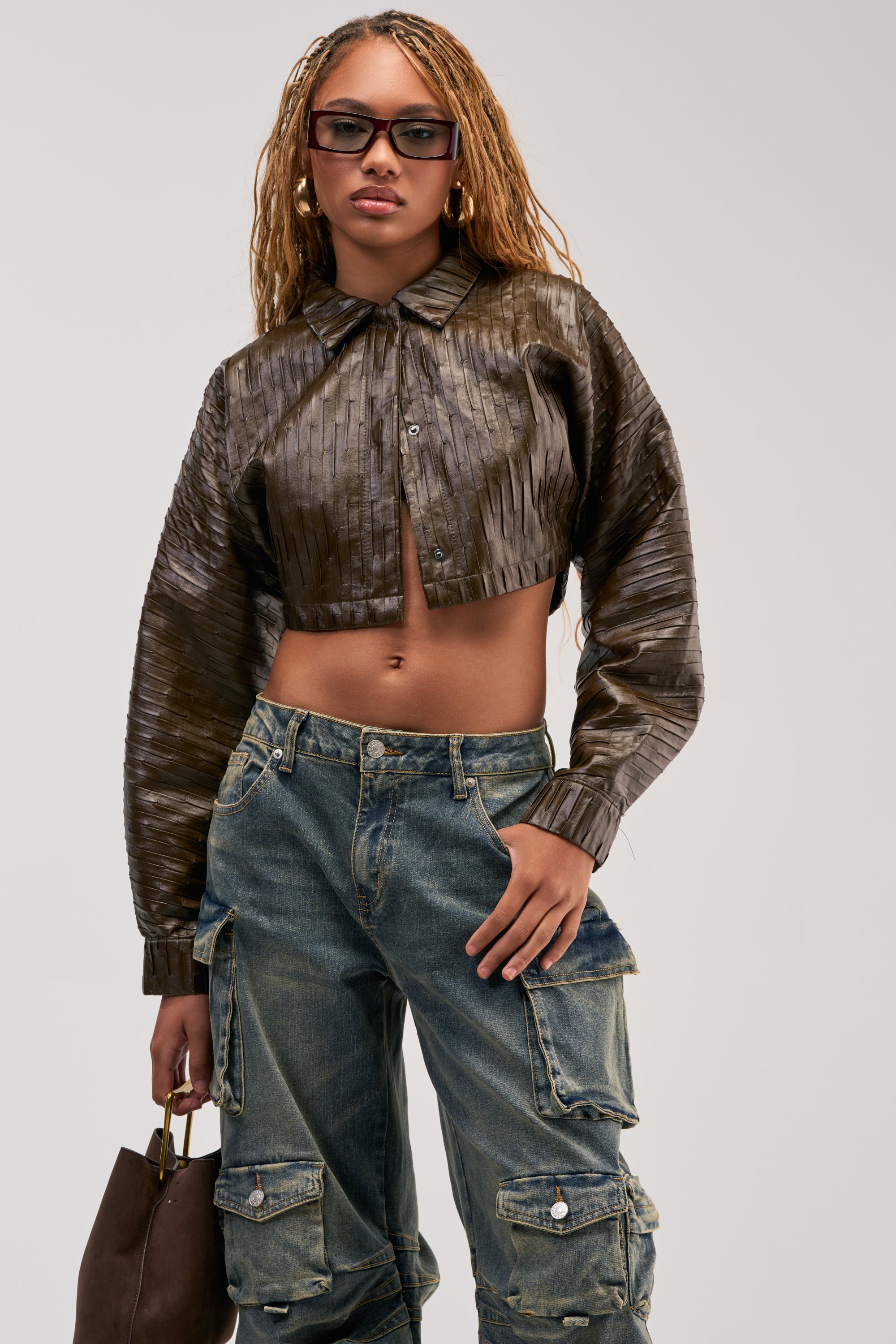 Woman with long braids wears sunglasses, the JACQUELINE TEXTURED PU CROPPED BUTTON DOWN in metallic brown, loose cargo jeans, and holds a brown handbag for a utility-chic look.
