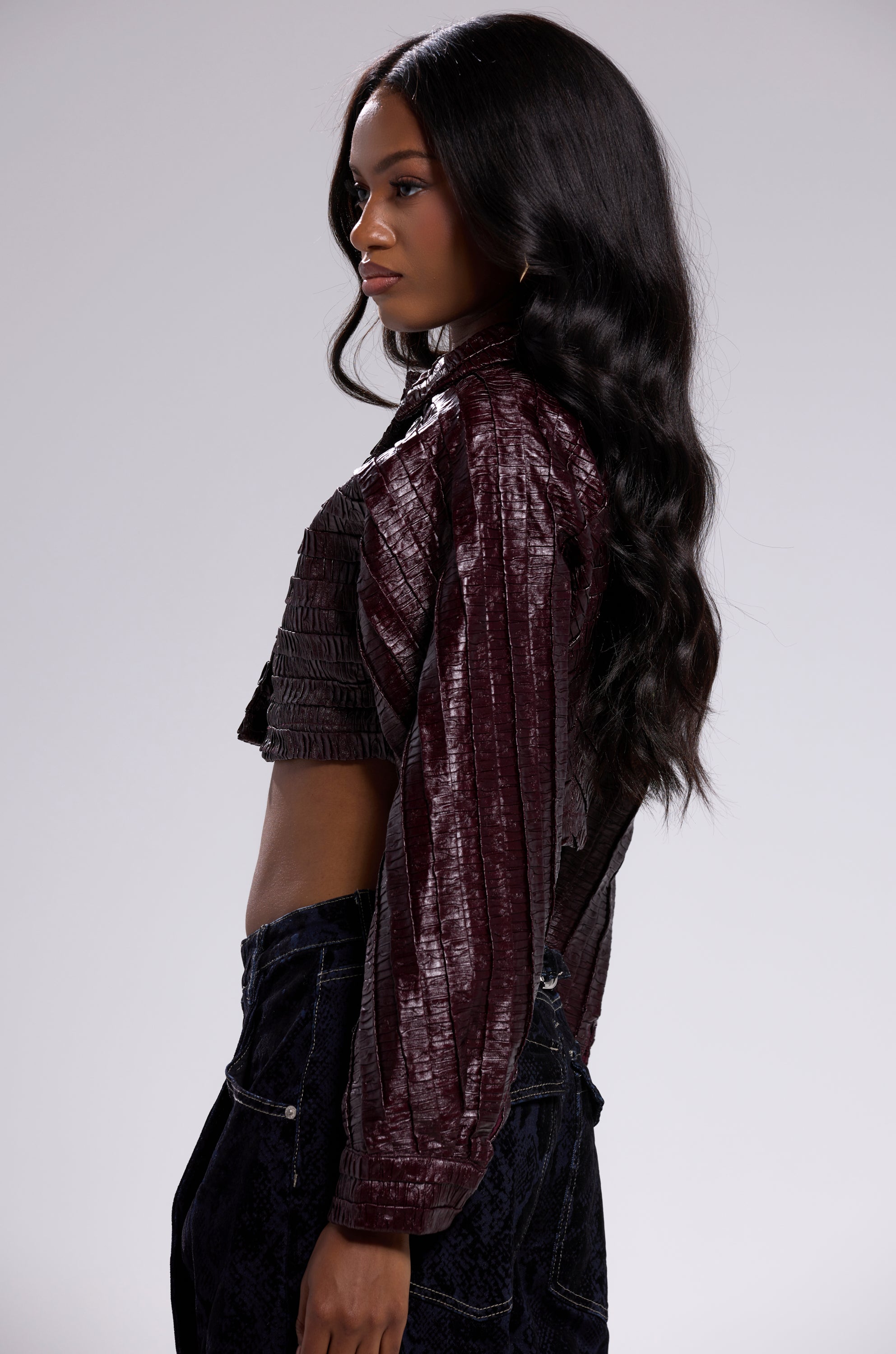 A woman with long wavy black hair stands in profile against a light gray background, wearing the JUST LIKE THAT BUTTON DOWN—a textured faux leather, dark burgundy, long-sleeve crop top—paired with black pants.