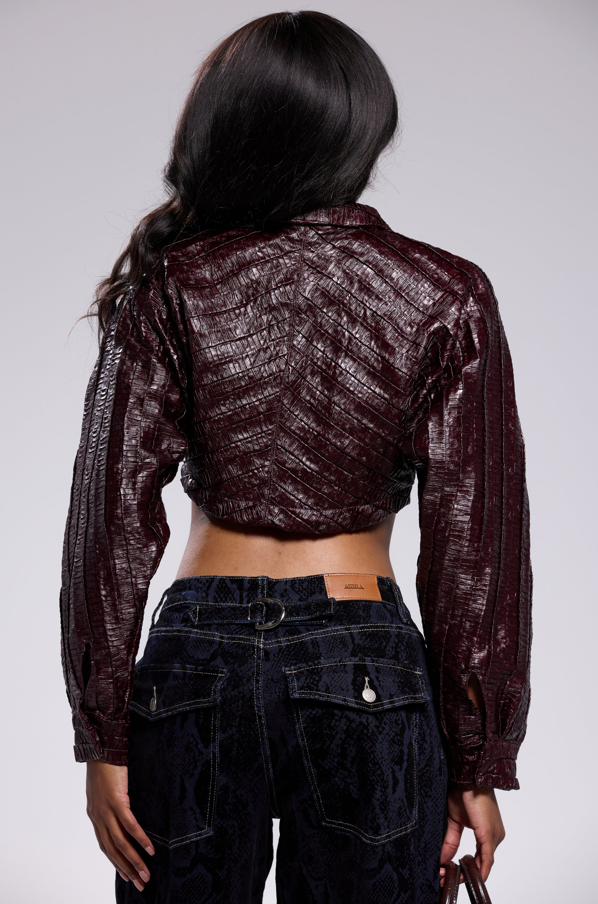 A woman stands facing away, wearing the JUST LIKE THAT BUTTON DOWN—a shiny dark burgundy cropped jacket—and high-waisted dark blue jeans with a subtle pattern. Her long, dark hair falls over her shoulder.