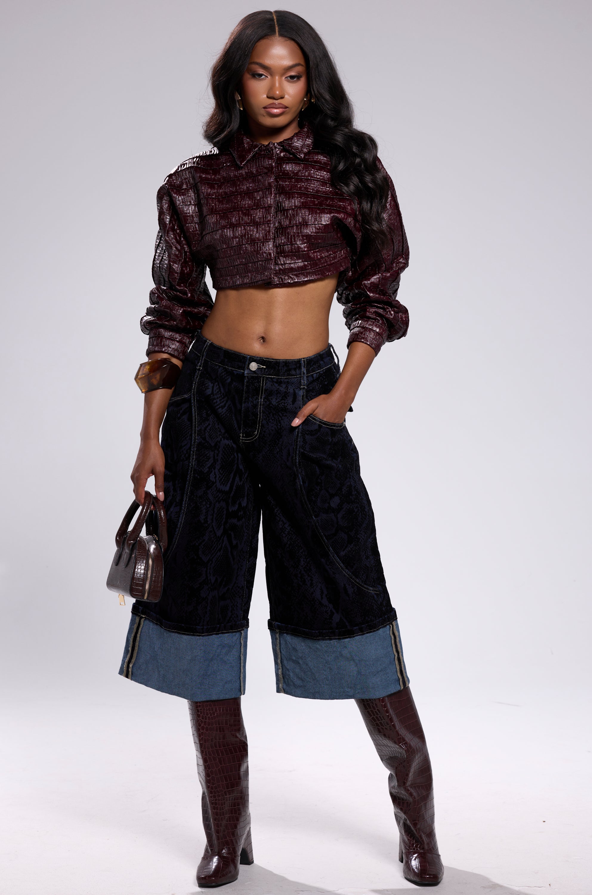 A woman poses confidently in the maroon JUST LIKE THAT BUTTON DOWN cropped shirt, dark wide-leg denim shorts with light cuffs, maroon knee-high boots, and holds a small brown handbag against a plain background.