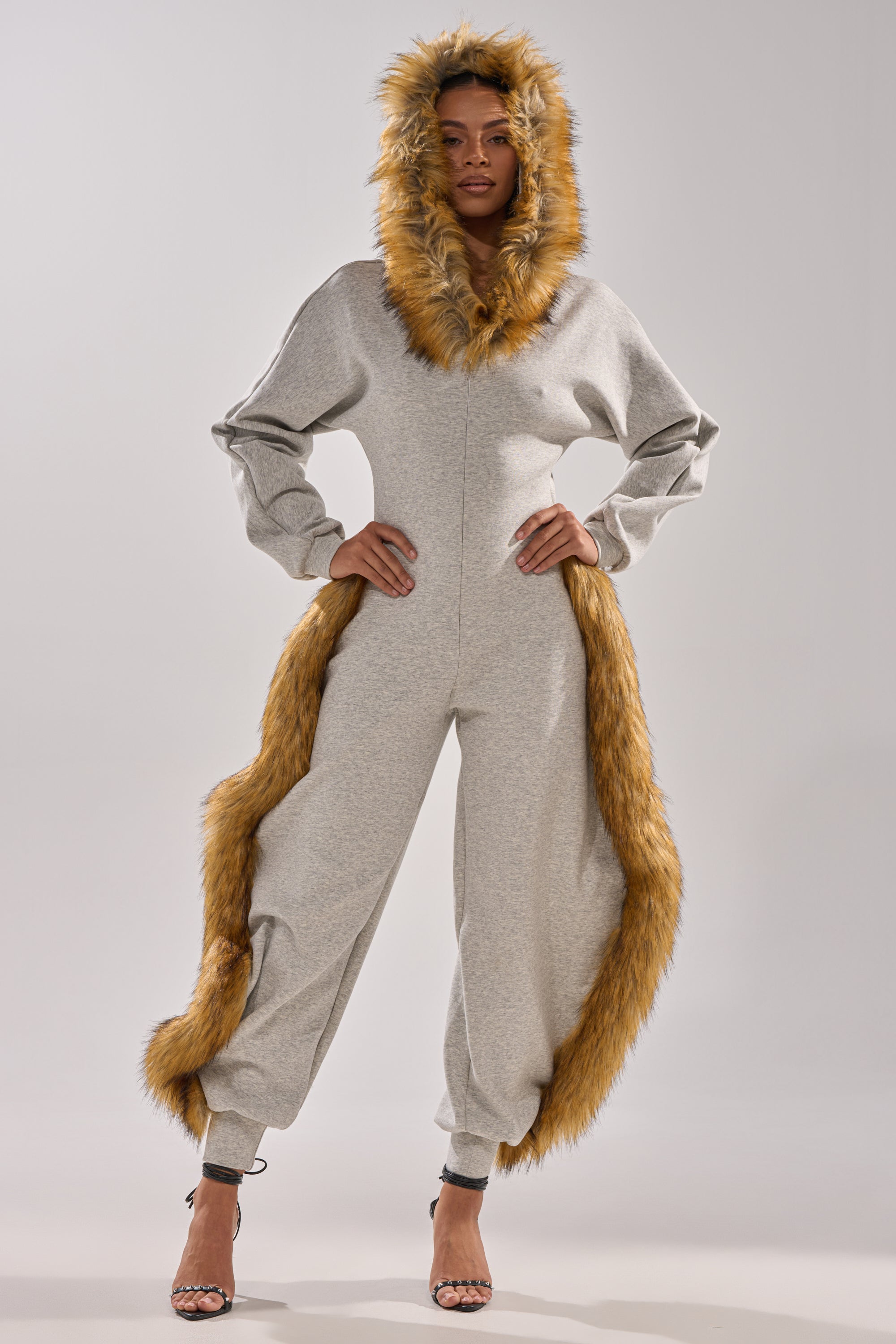 A woman stands confidently in the YOU WISH JUMPSUIT WITH FUR ACCENTS, a hooded one-piece with plush faux fur trim and a long tail, paired with black high-heeled sandals for a chic, animal-inspired look.