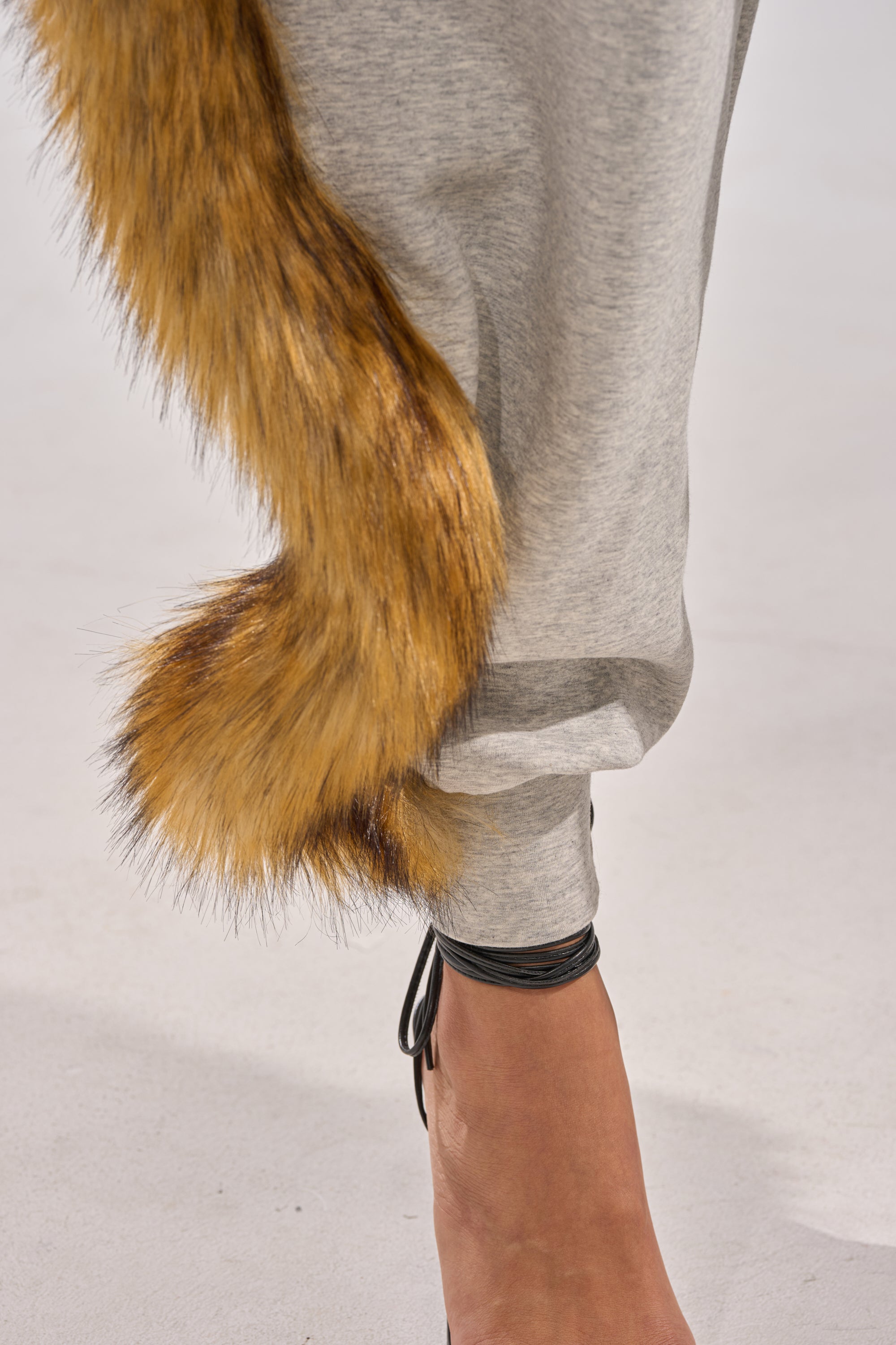 Close-up of a person wearing the YOU WISH JUMPSUIT WITH FUR ACCENTS in light gray, styled with a brown faux fur tail accessory and black strappy high heels on a light-colored surface.