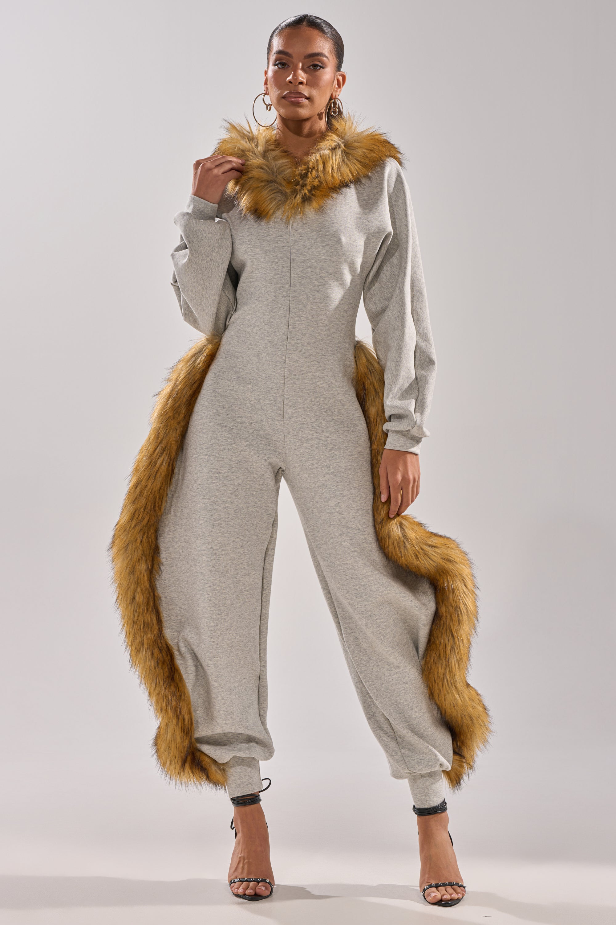 A woman models the YOU WISH JUMPSUIT WITH FUR ACCENTS in light gray, featuring long sleeves and brown faux fur trim on the collar and sides, styled with high-heeled sandals against a simple light background.
