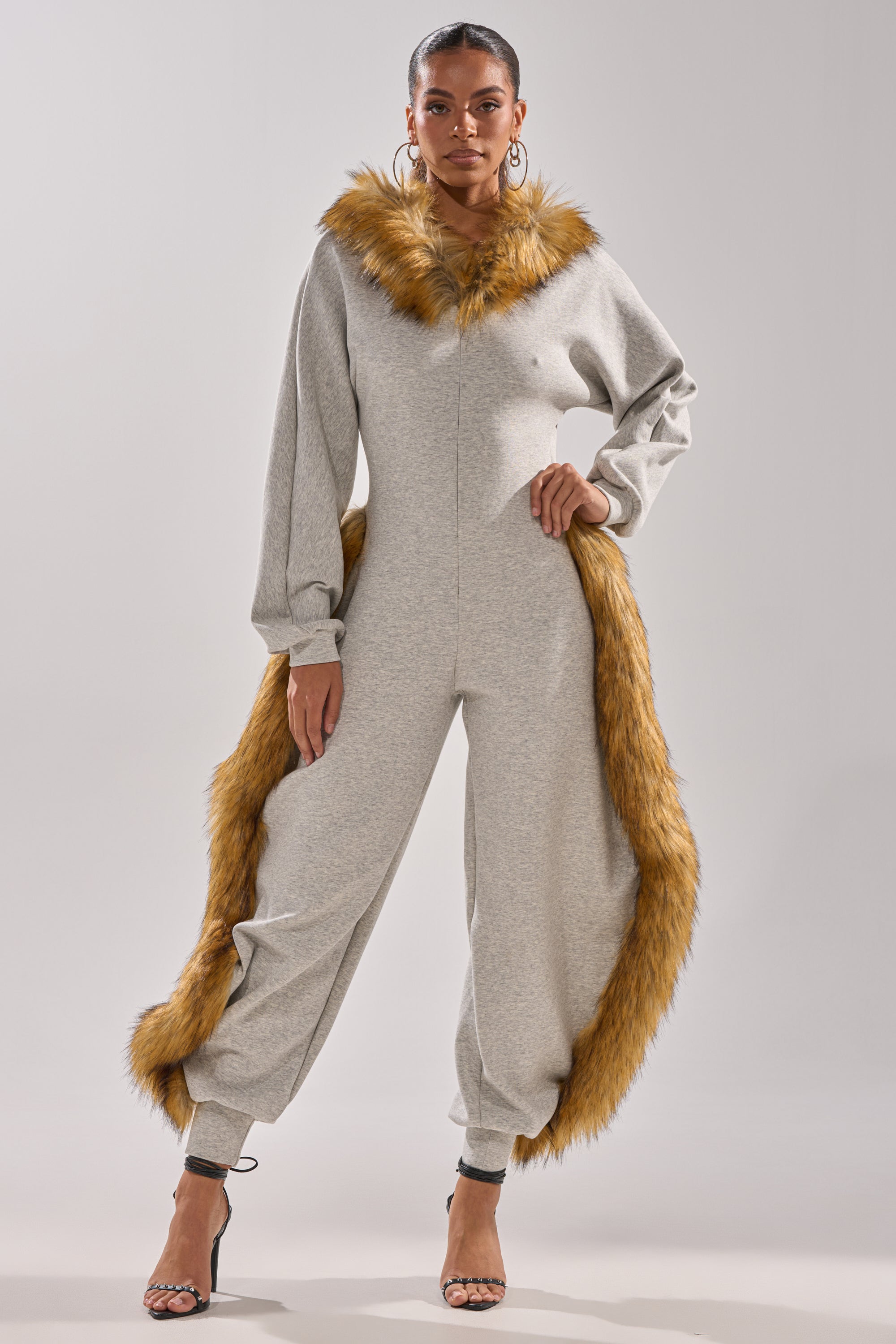 A woman stands confidently in the YOU WISH JUMPSUIT WITH FUR ACCENTS, featuring plush faux fur with brown trim on the hood and leg, styled with hoop earrings and black heels against a plain light background.