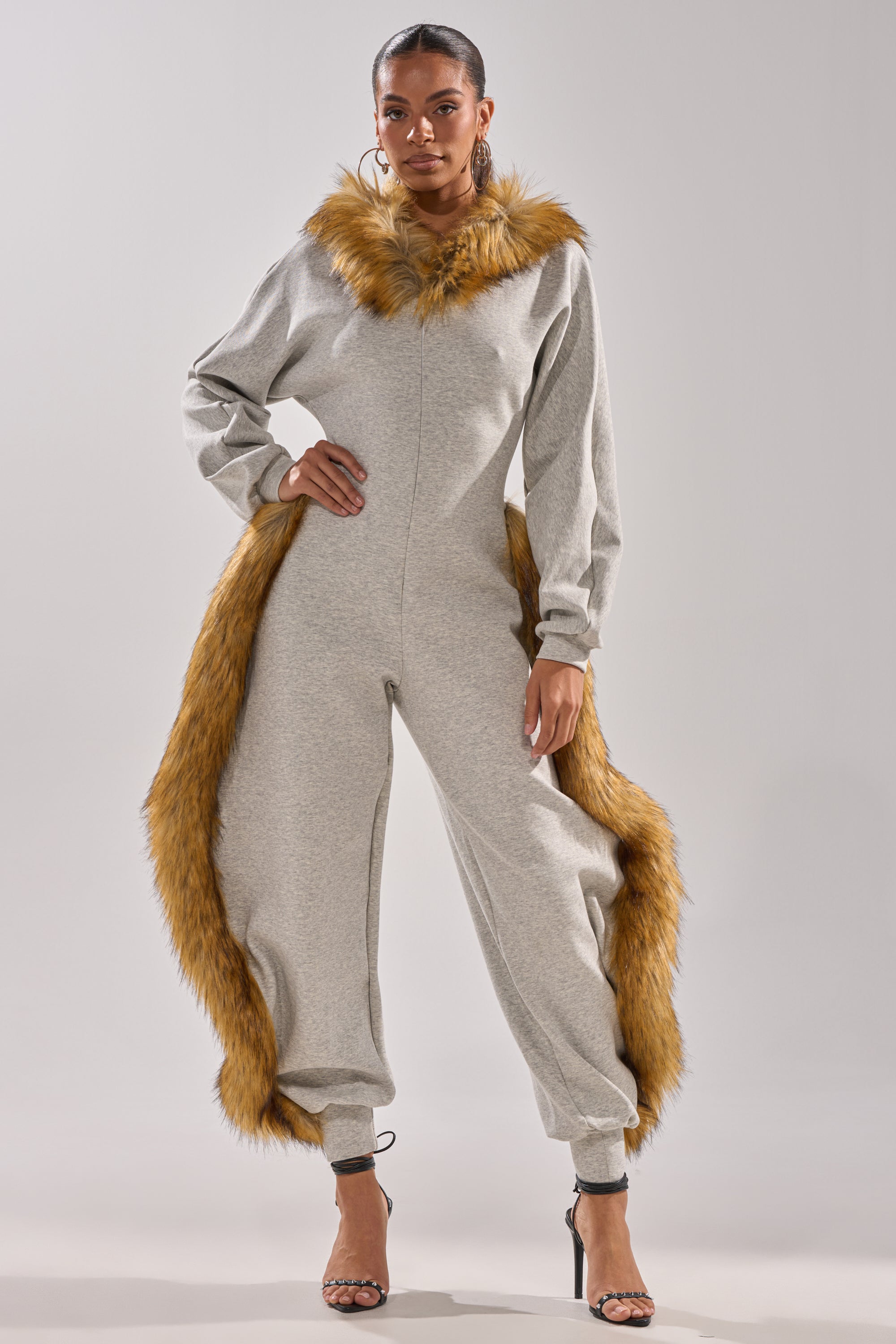 A woman models the YOU WISH JUMPSUIT WITH FUR ACCENTS, featuring bold brown faux fur around the collar, hips, and ankles. She pairs it with black high-heeled sandals against a simple light background.