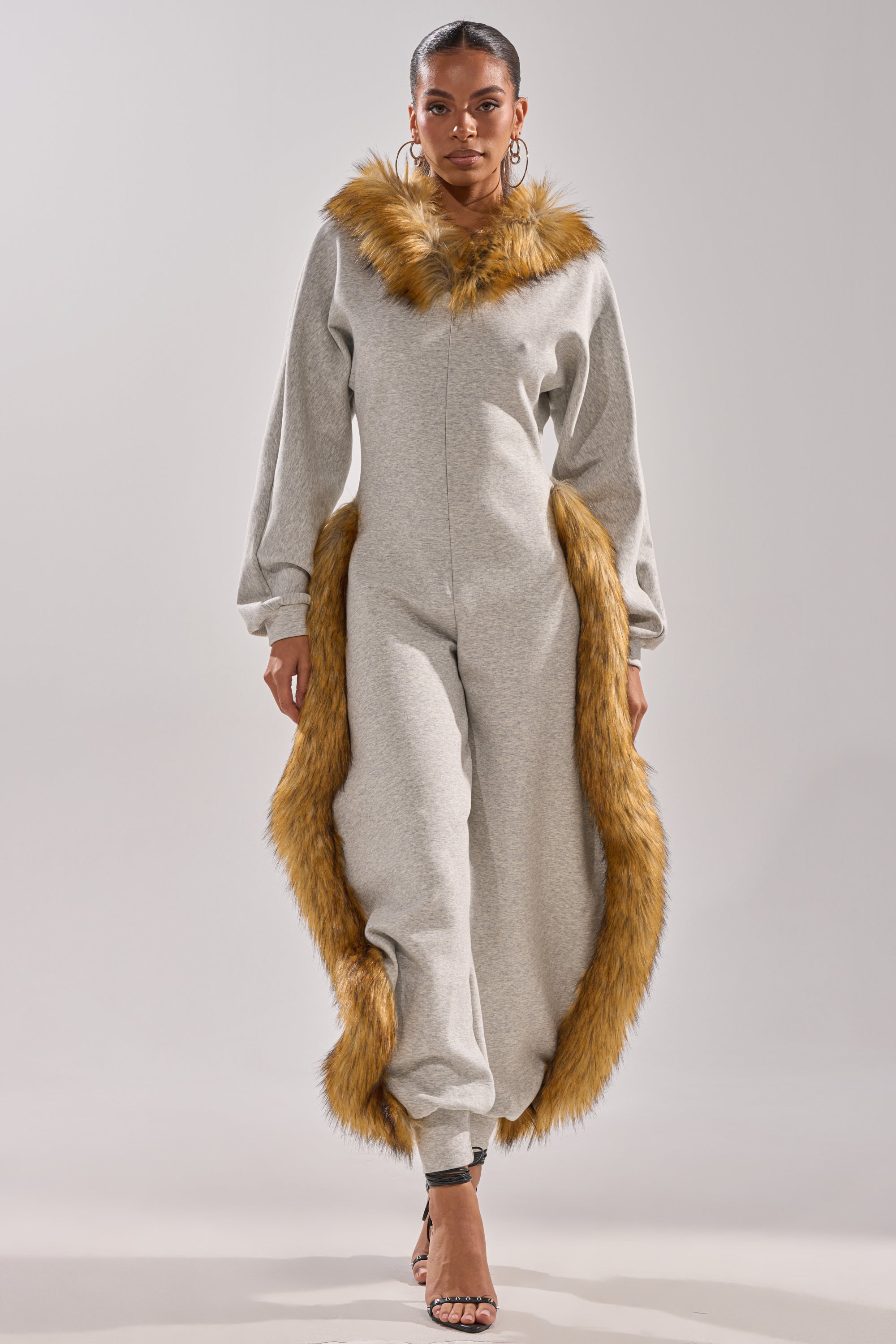 A woman wears the YOU WISH JUMPSUIT WITH FUR ACCENTS, featuring wide sleeves and brown faux fur trim on the collar and sides, paired with hoop earrings and high-heeled sandals, standing against a plain background.