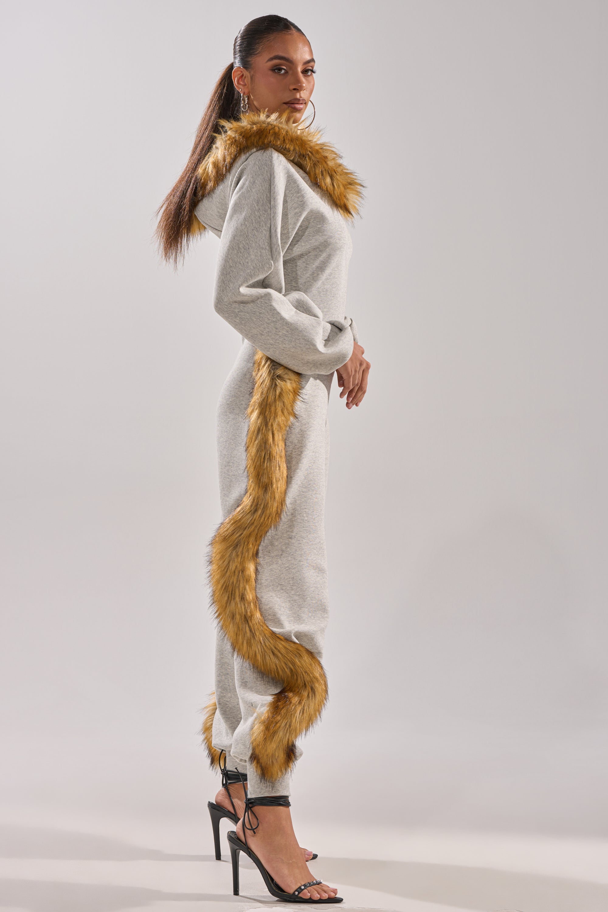 A woman poses in profile against a light background wearing the YOU WISH JUMPSUIT WITH FUR ACCENTS—a grey, hooded one-piece with fur trim along the hood, shoulders, and sides—styled with black high-heeled sandals.
