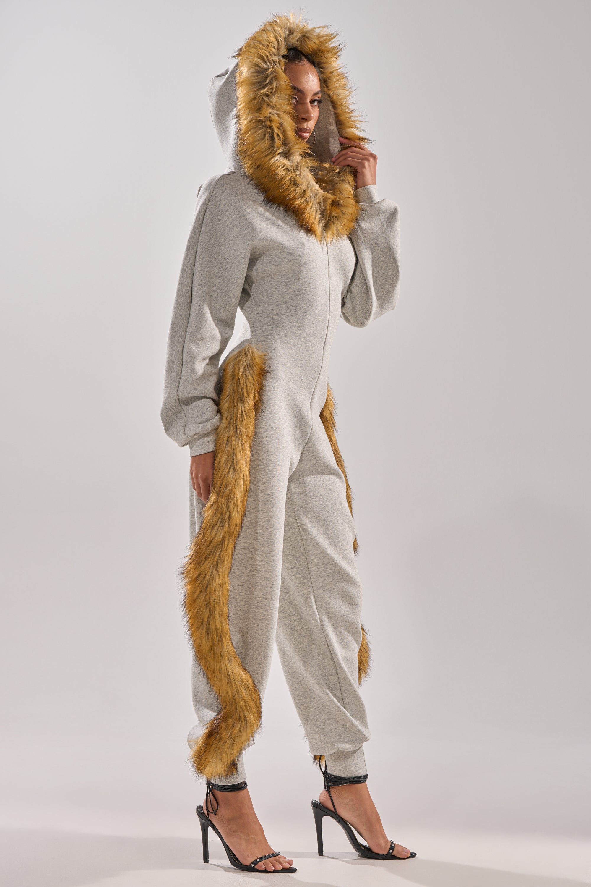 Model in profile wears the YOU WISH JUMPSUIT WITH FUR ACCENTS—light gray faux fur with brown trim on hood, sides, and cuffs. Black high-heeled sandals finish this chic look against a plain background.