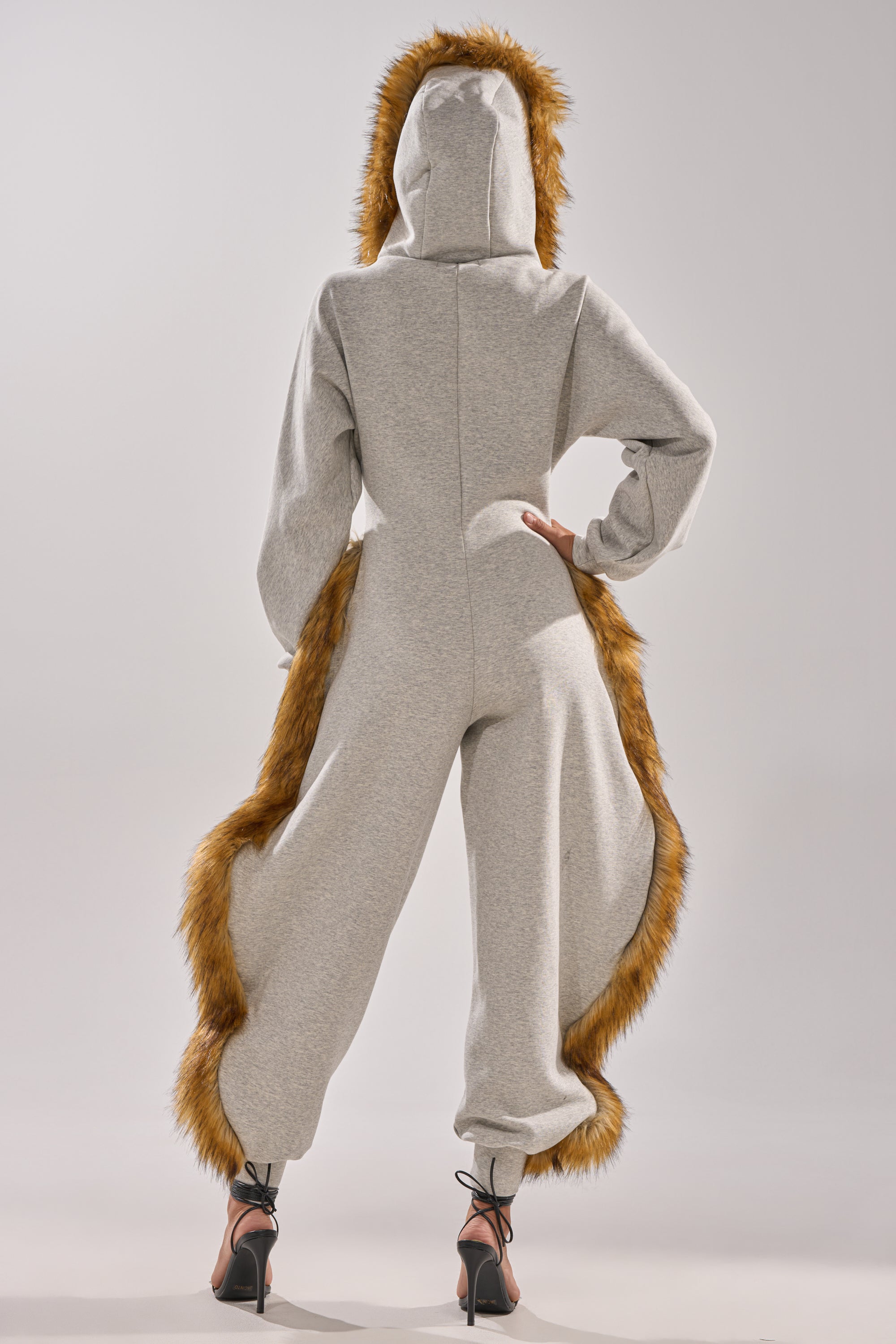 Person shown from behind wearing the YOU WISH JUMPSUIT WITH FUR ACCENTS—features a plush faux fur hood and tail-inspired trim—paired with black high heels, set against a plain light background.