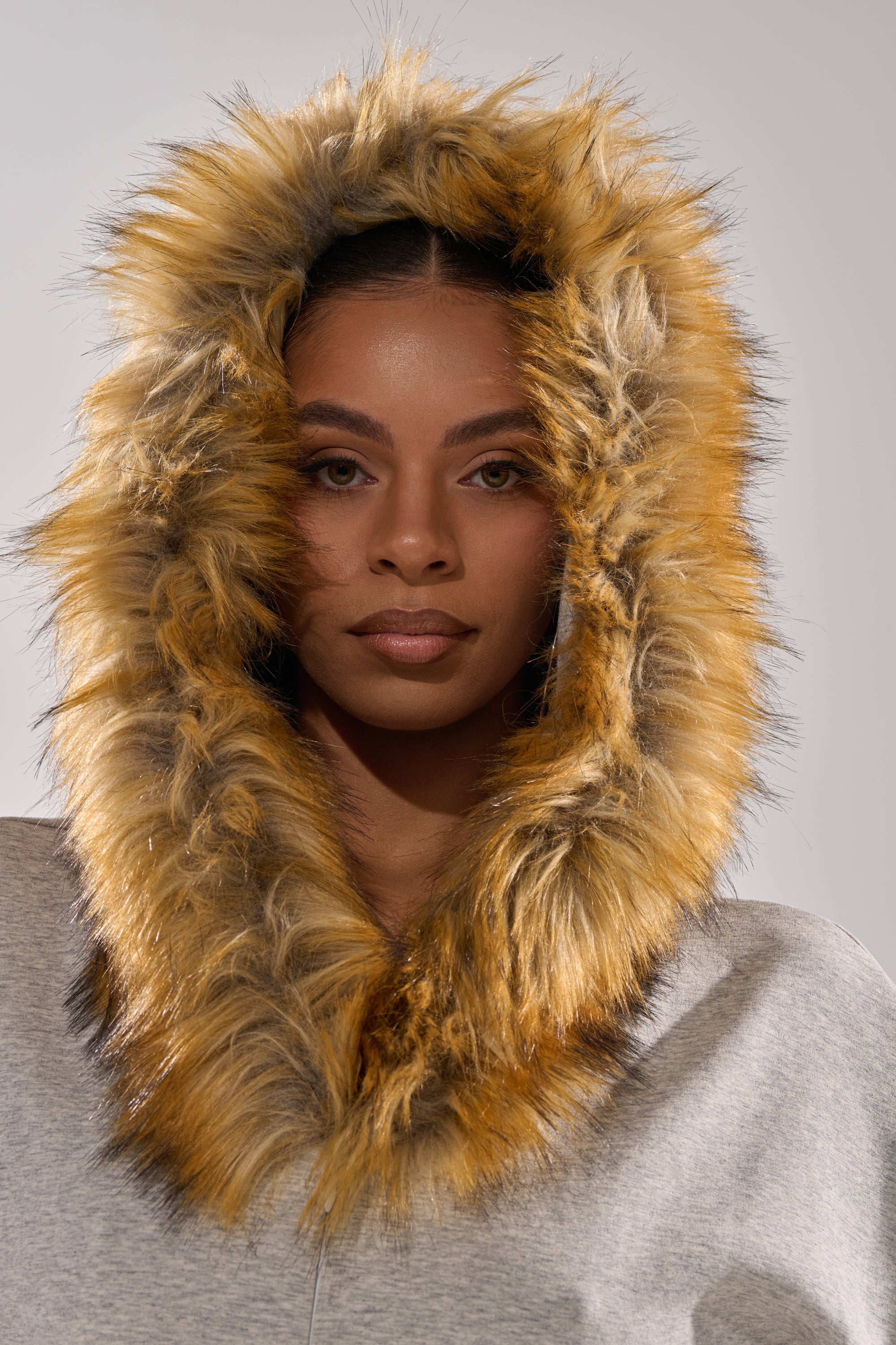 A woman models the YOU WISH JUMPSUIT WITH FUR ACCENTS, featuring a gray hood with wide, fluffy brown and yellow faux fur trim framing her face as she looks at the camera against a plain background.