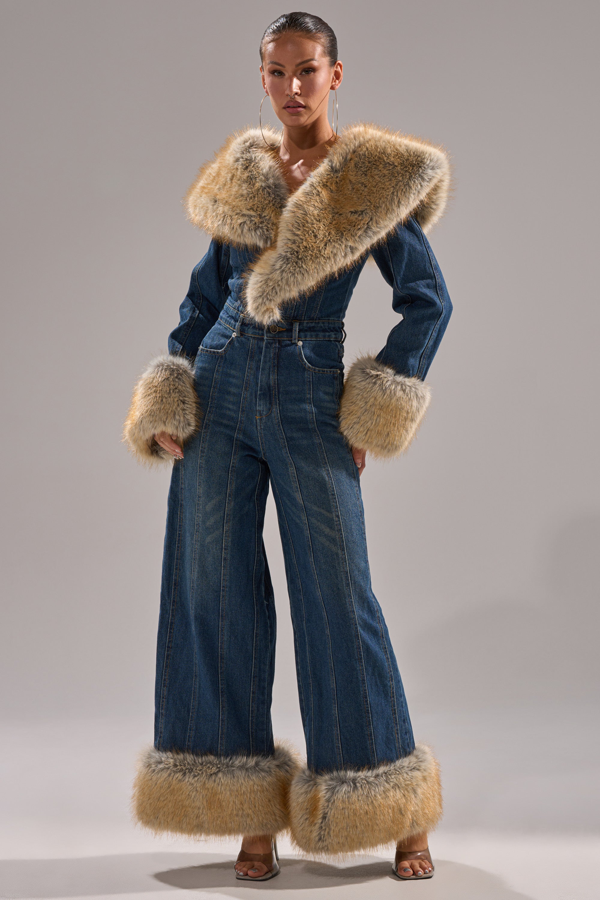 A person models the LENNOX DENIM JUMPSUIT WITH FUR ACCENTS, featuring a wide-leg cut and tan faux fur trim on the collar, cuffs, and ankles, set against a plain gray background.