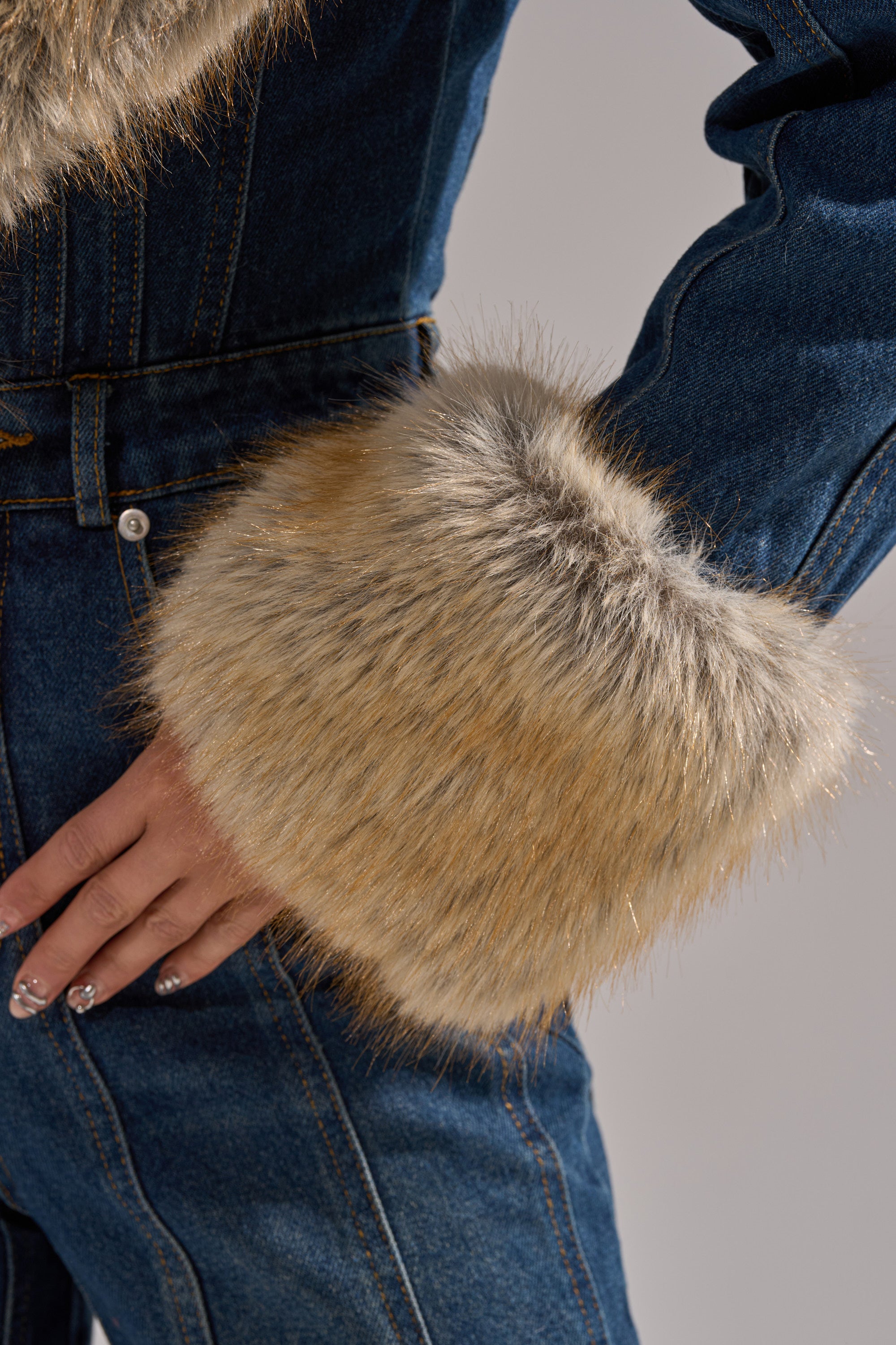 A close-up shows a hand on the hip, wearing the LENNOX DENIM JUMPSUIT WITH FUR ACCENTS. The wide-leg style and plush faux fur cuffs bring flair, while manicured nails and a neutral background complete this chic look.