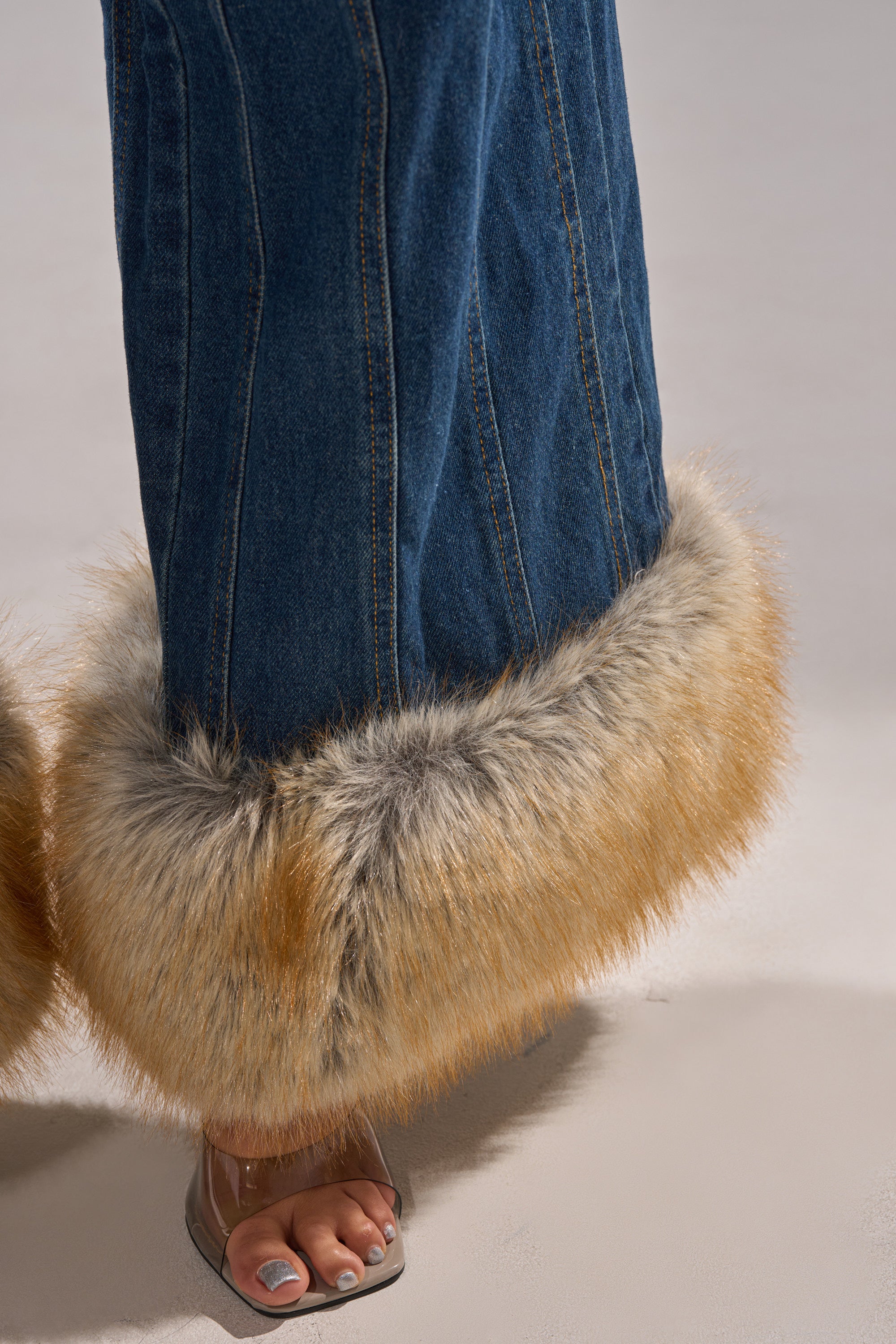 A close-up of a person's lower legs showcases the LENNOX DENIM JUMPSUIT WITH FUR ACCENTS, featuring wide-leg blue denim and thick tan and grey faux fur cuffs, paired with clear open-toe heels and white-painted toenails.