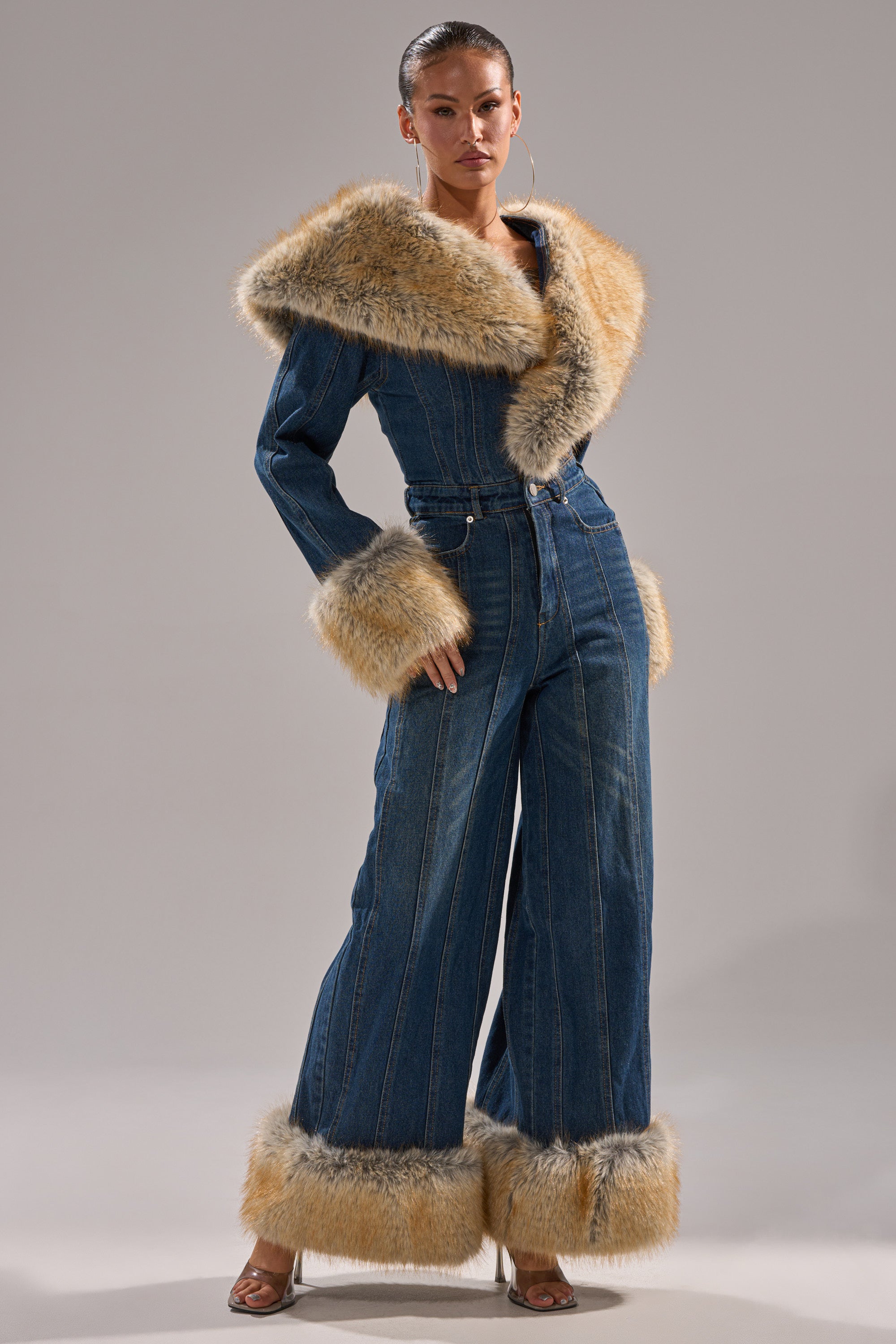 A woman stands confidently in the LENNOX DENIM JUMPSUIT WITH FUR ACCENTS, showcasing its wide-leg silhouette and tan faux fur trim on the collar, cuffs, and hem against a plain background.
