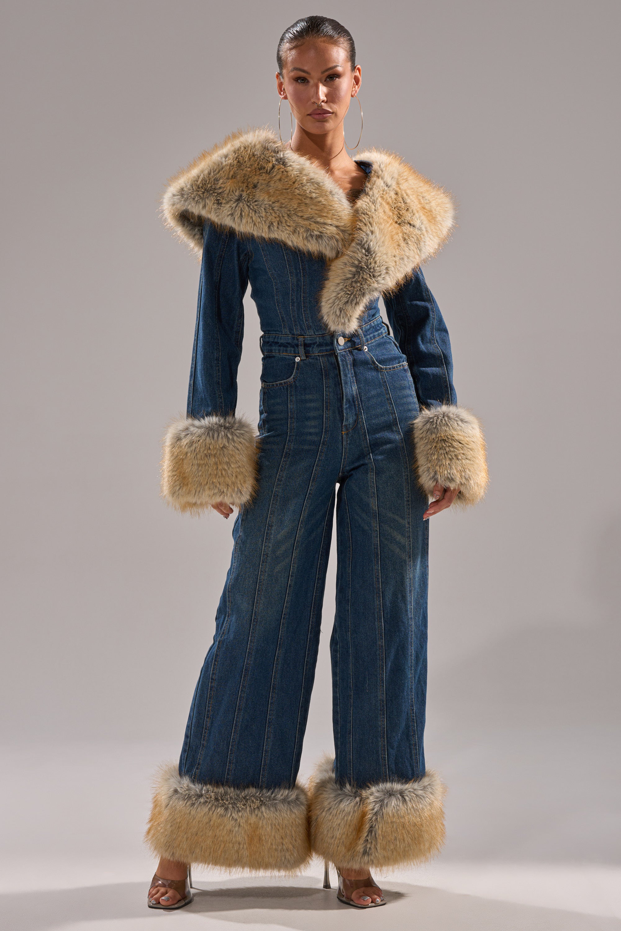A woman exudes confidence in the LENNOX DENIM JUMPSUIT WITH FUR ACCENTS, featuring wide legs and faux fur details on the collar, cuffs, and hems, styled with slicked-back hair, hoop earrings, and heeled sandals.