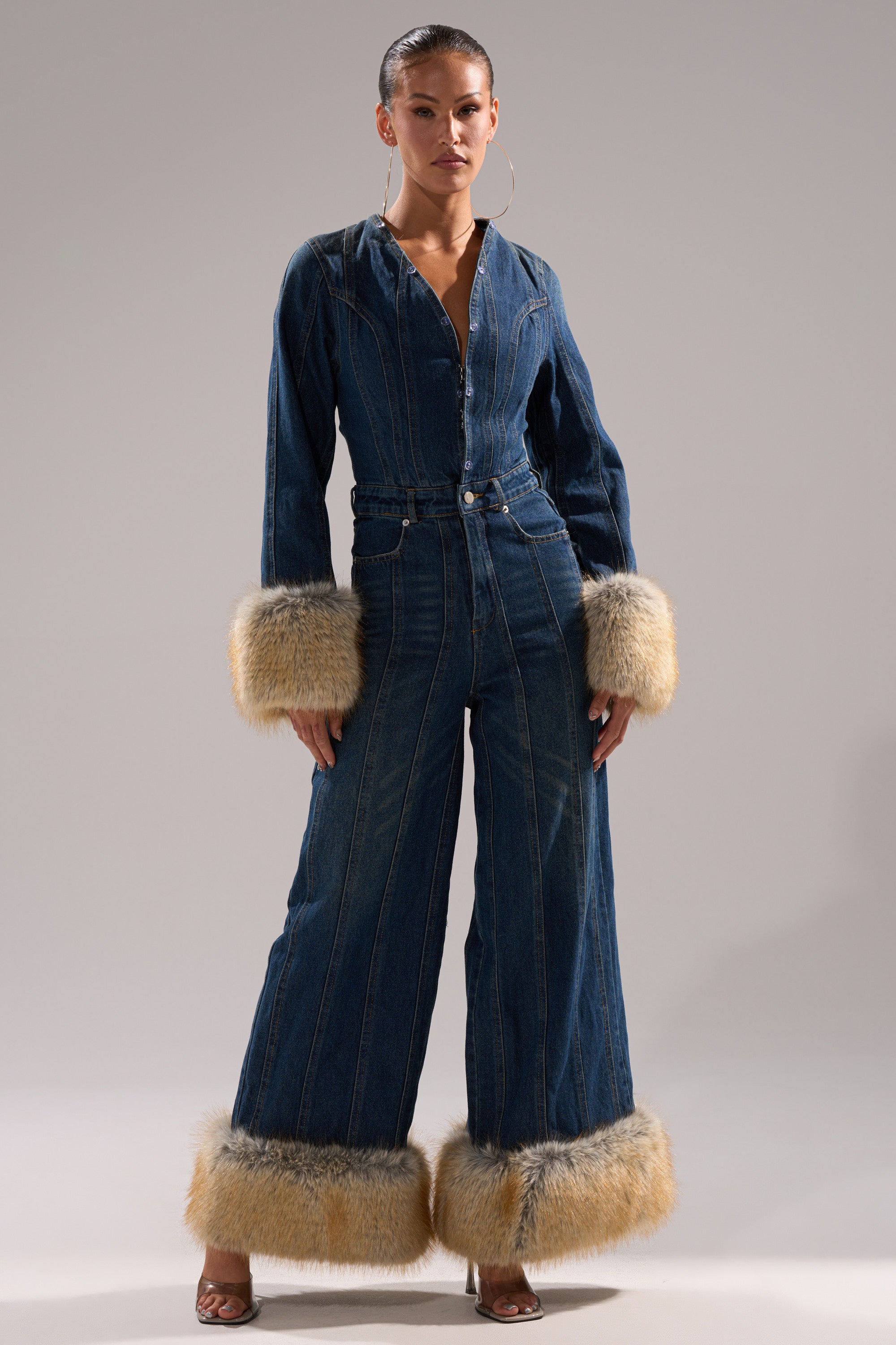 A woman poses confidently in the LENNOX DENIM JUMPSUIT WITH FUR ACCENTS, featuring a wide-leg silhouette and long sleeves with tan faux fur at the cuffs and hem, styled with hoop earrings and heeled sandals against a plain background.