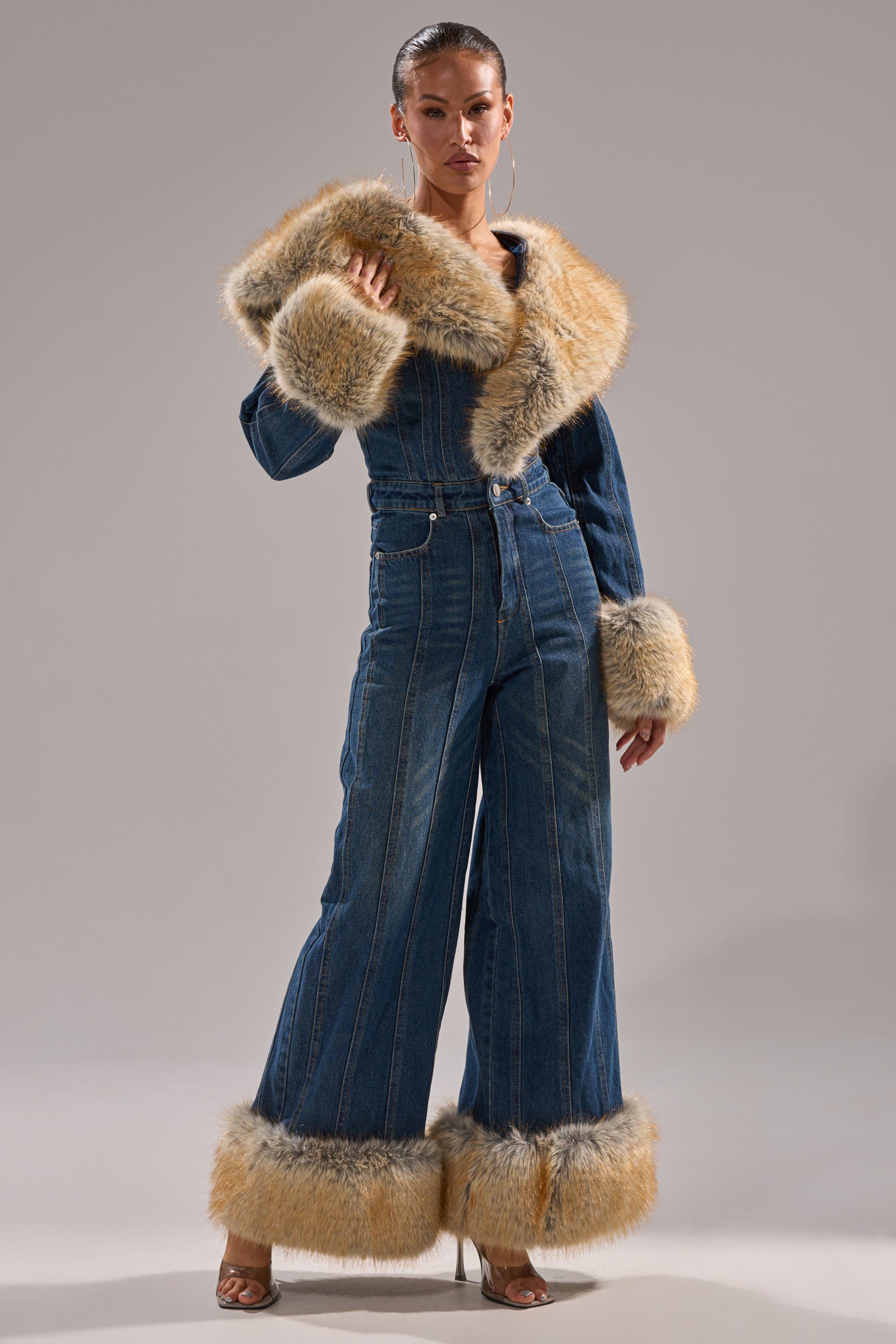 A woman models the LENNOX DENIM JUMPSUIT WITH FUR ACCENTS, featuring a wide-leg silhouette and tan faux fur trim on the collar, cuffs, and hems, paired with hoop earrings and heeled sandals against a plain gray background.