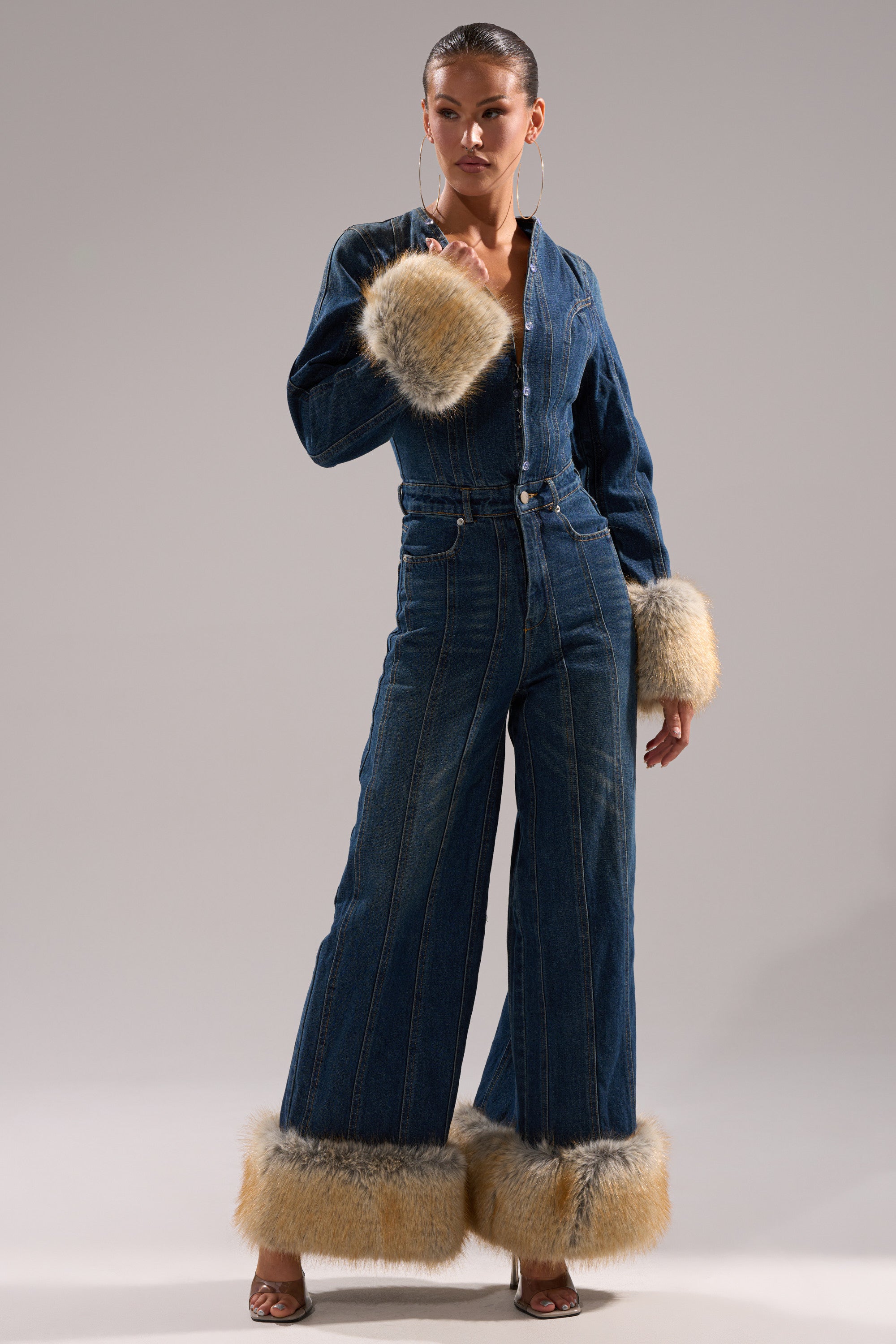 A woman stands confidently in the LENNOX DENIM JUMPSUIT WITH FUR ACCENTS, showcasing its wide-leg silhouette and fur-trimmed cuffs and hems, paired with hoop earrings and heeled sandals against a plain background.