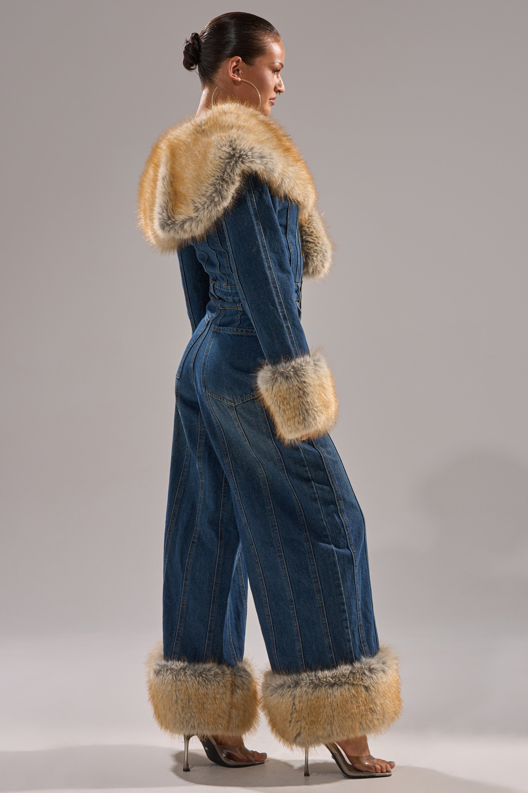 A person stands sideways in the LENNOX DENIM JUMPSUIT WITH FUR ACCENTS, featuring tan faux fur on the hood, cuffs, and pant legs. The tailored fit flows into a wide-leg silhouette, styled with high-heeled sandals.