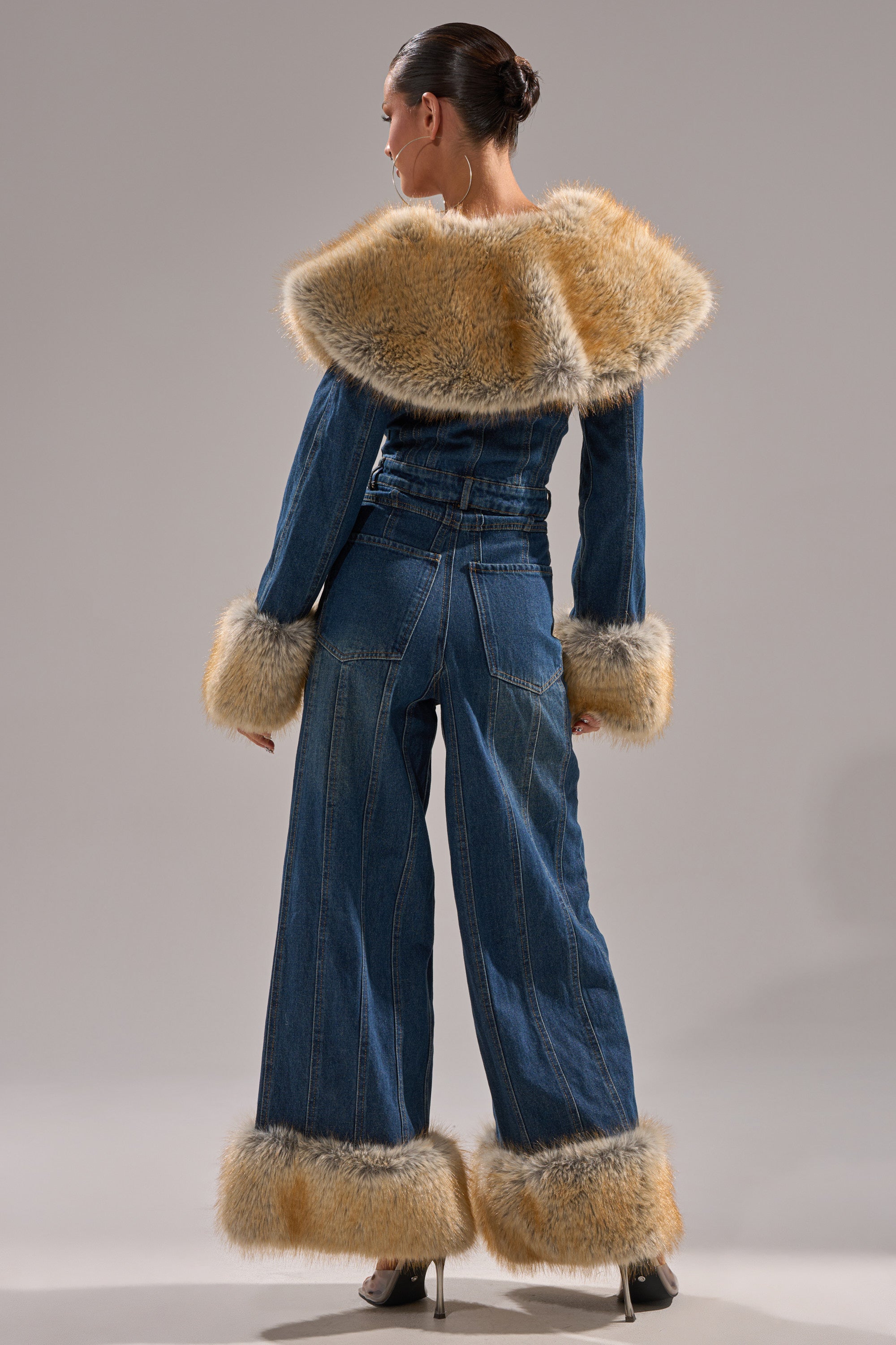 A woman models the LENNOX DENIM JUMPSUIT WITH FUR ACCENTS, featuring a wide-leg silhouette and tan faux fur trim on the collar, cuffs, and pant legs. She styles her hair in a sleek bun and pairs the look with high heels.